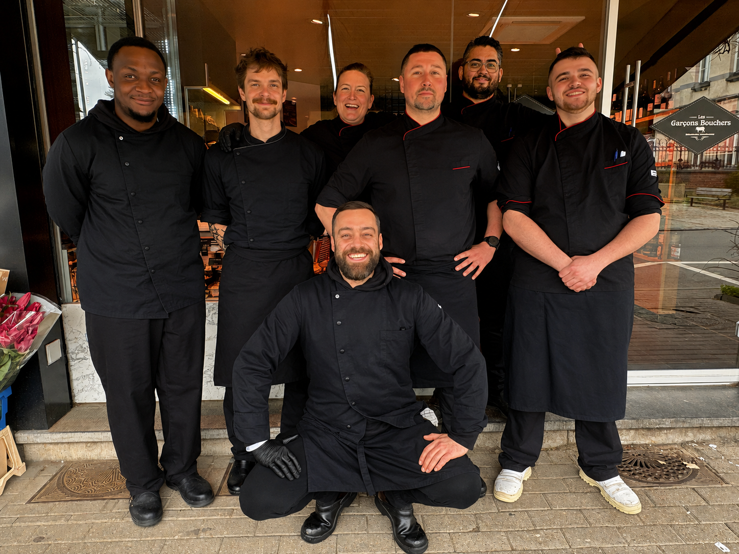 Groupe de sept chefs en uniforme noir posant devant un restaurant.