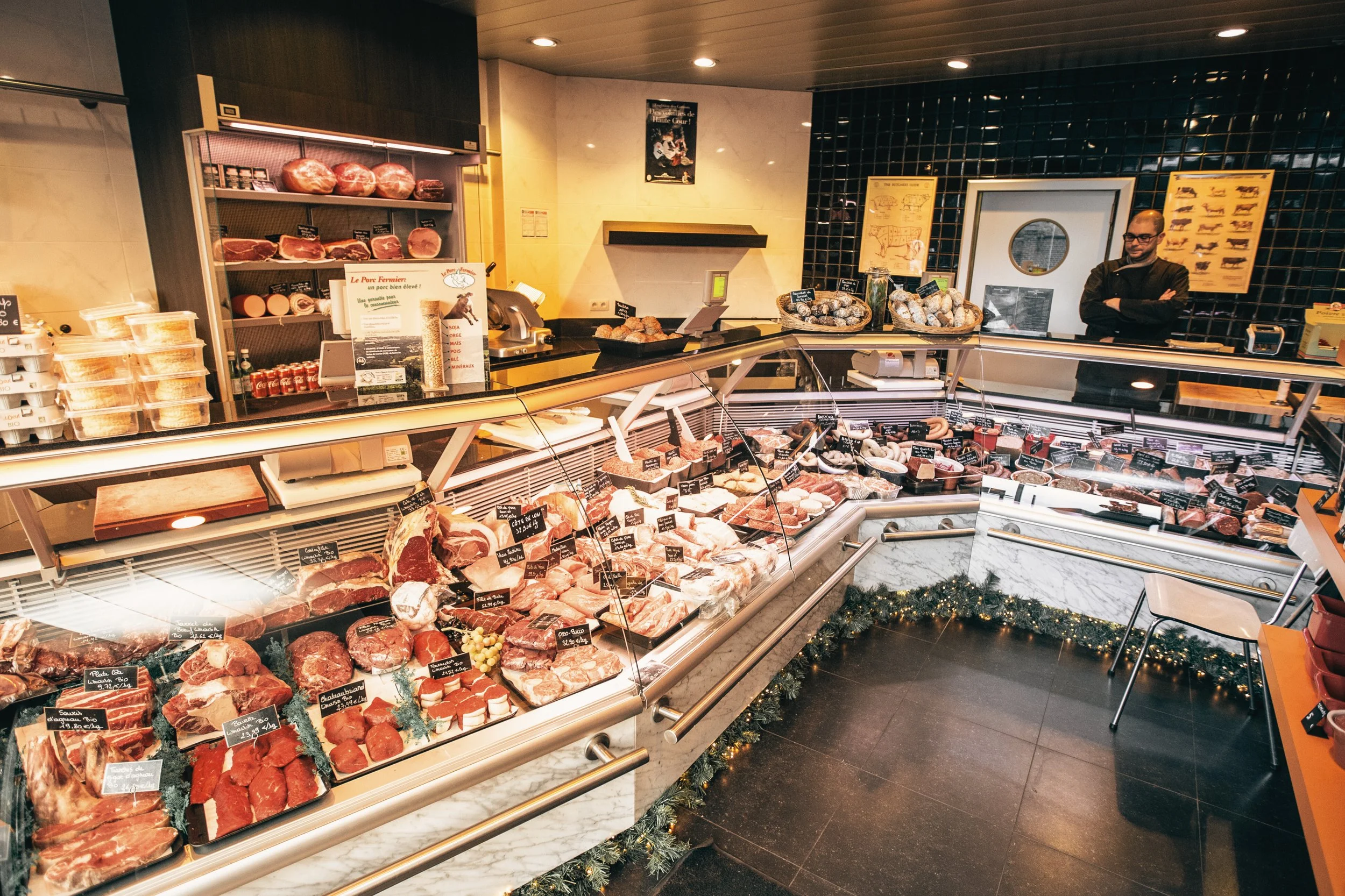 Une boucherie avec un comptoir rempli de diverses viandes, et un homme derrière le comptoir. La vitrine contient différents morceaux de viande, avec des étiquettes indiquant les prix et types. En arrière-plan, des posters illustrant des animaux et des coupes de viande sont affichés.