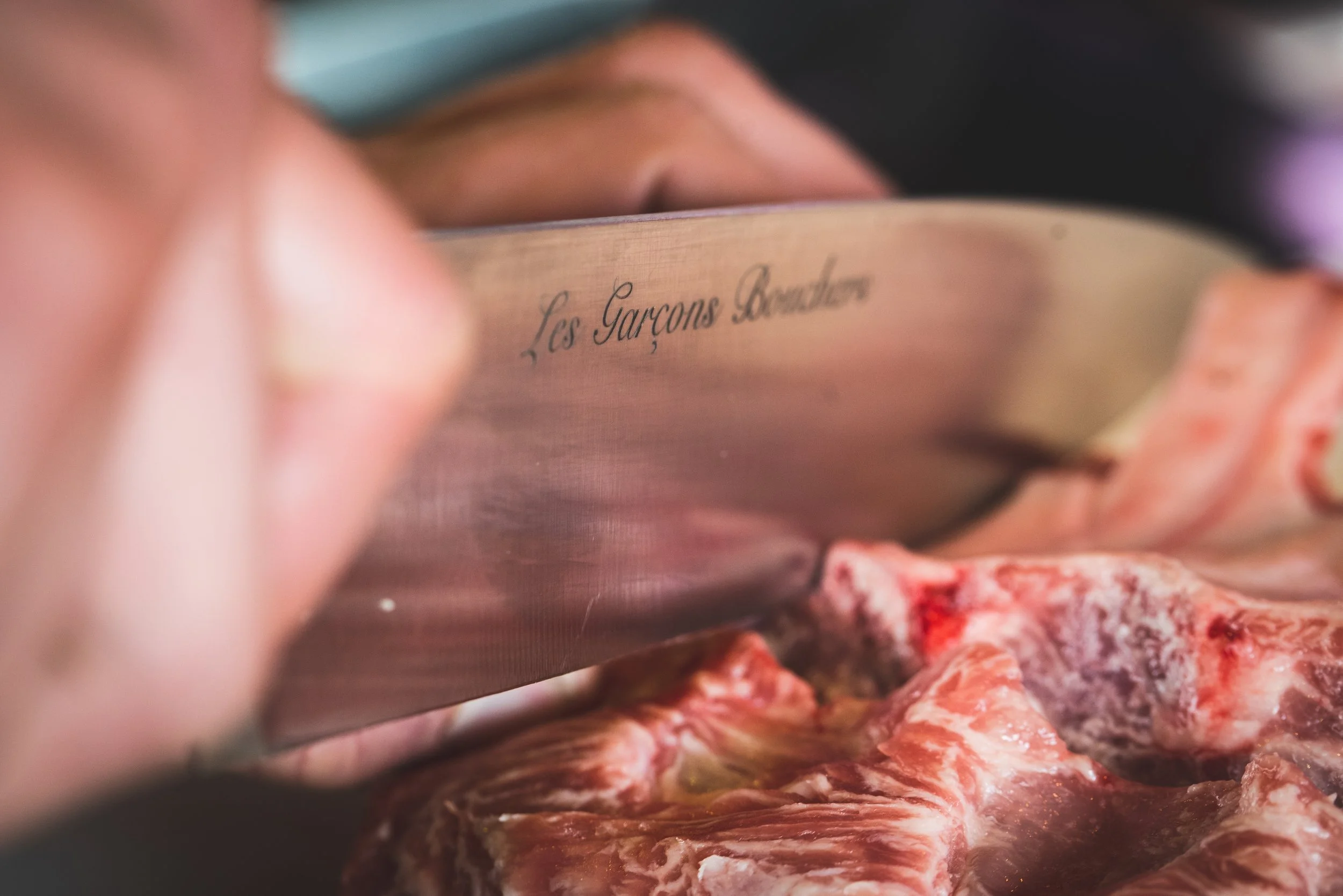 Une main utilisant un couteau pour couper de la viande rouge, avec un texte écrit en cursive sur le côté qui dit 'Les Garçons Bouchers'.