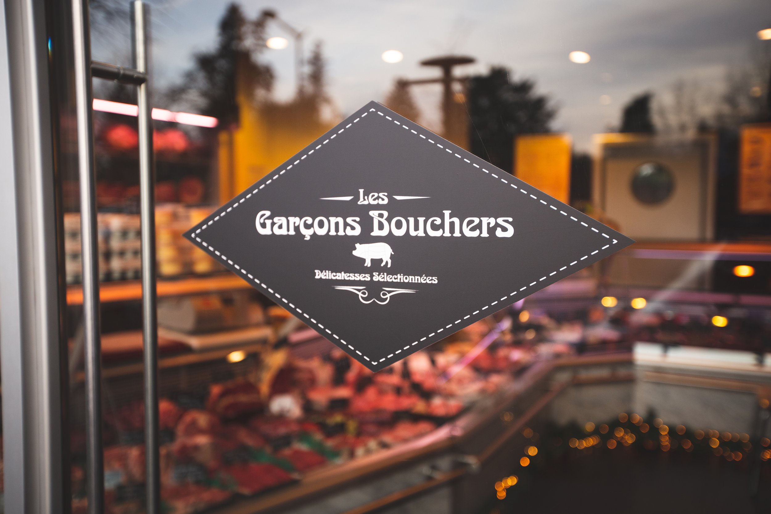 Une vitre de magasin de boucherie avec un panneau indiquant « Les Garçons Bouchers », un logo de cochon, et la mention « Délicatesses Sélectionnées », à l'extérieur du magasin avec une façade de produits frais en arrière-plan.