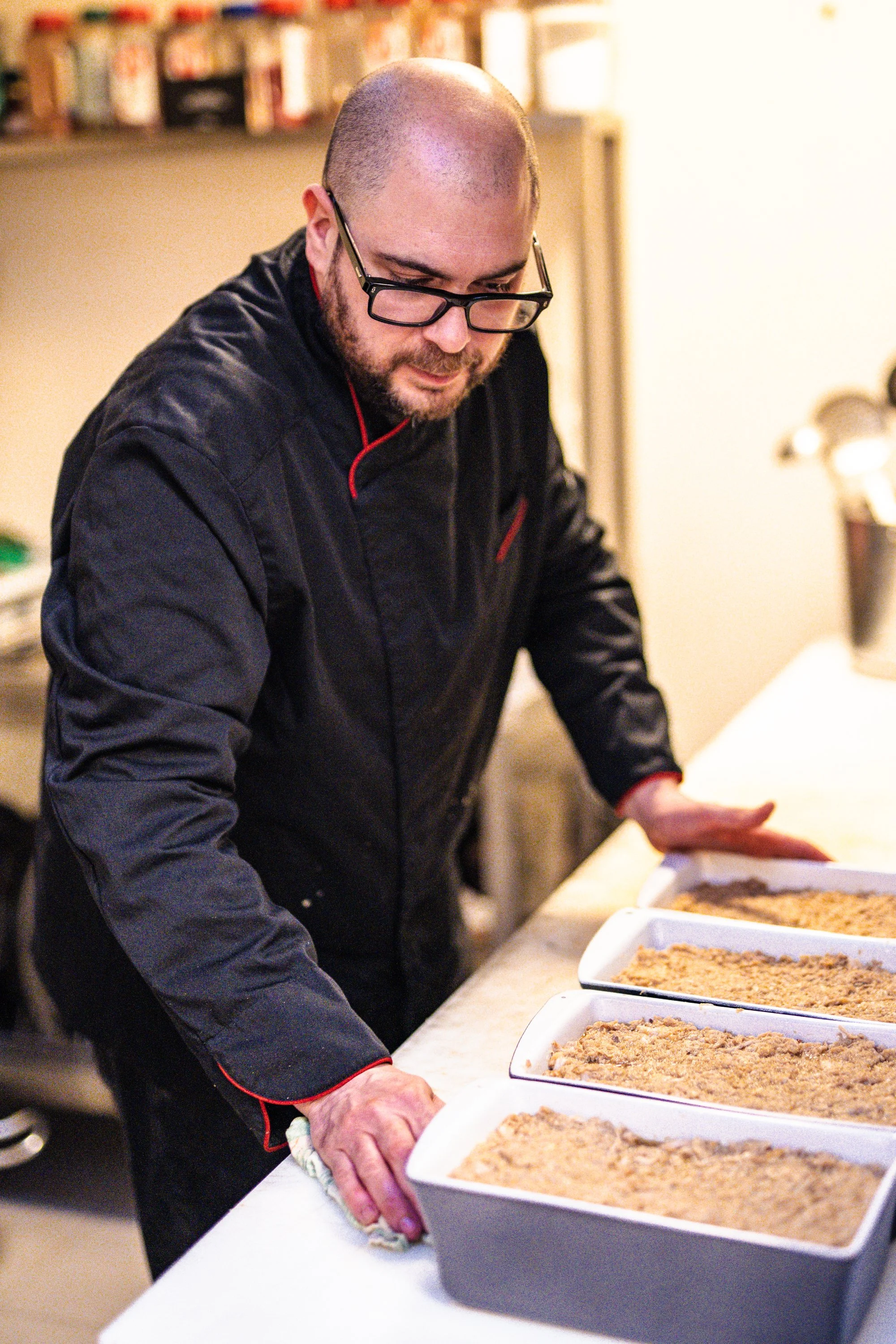 Un homme portant des lunettes et un tablier noir en train de préparer des plats en cuisine, avec plusieurs  préparations artisanales devant lui