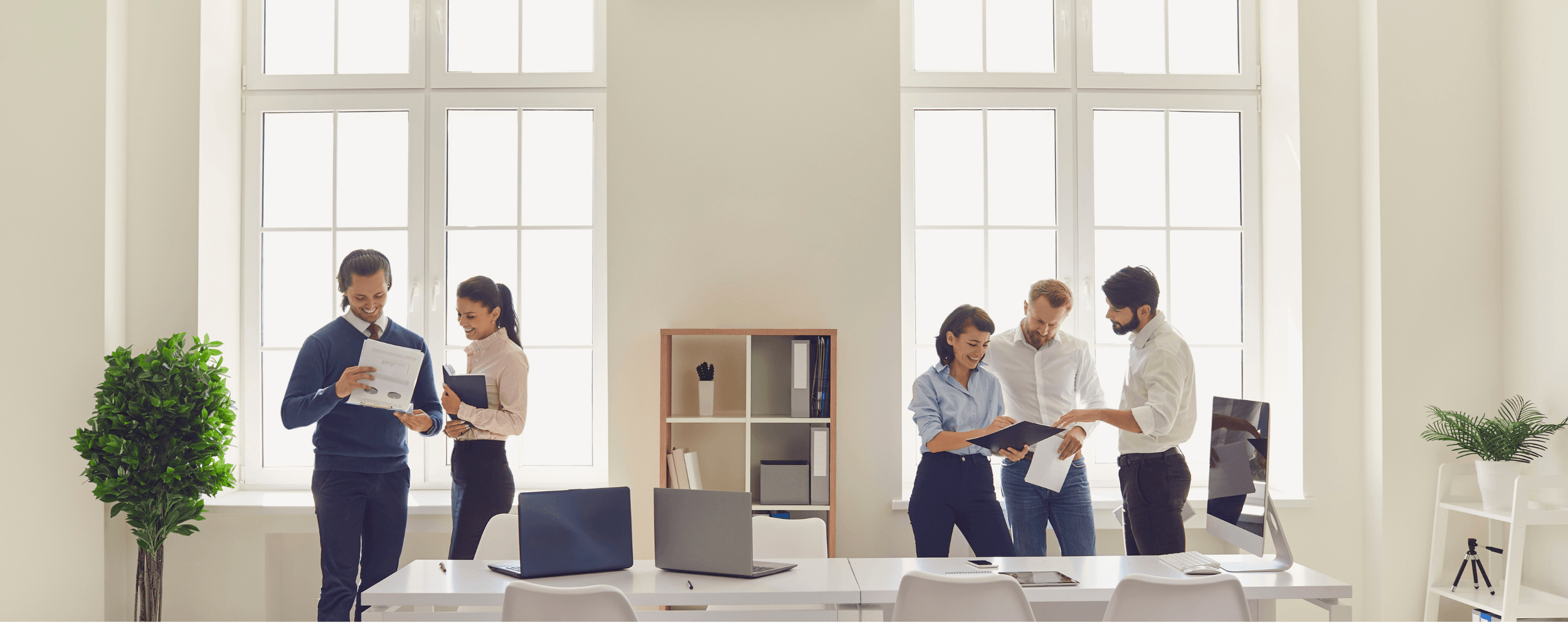 Group of six diverse professionals in a bright, modern office with large windows, working and discussing documents.