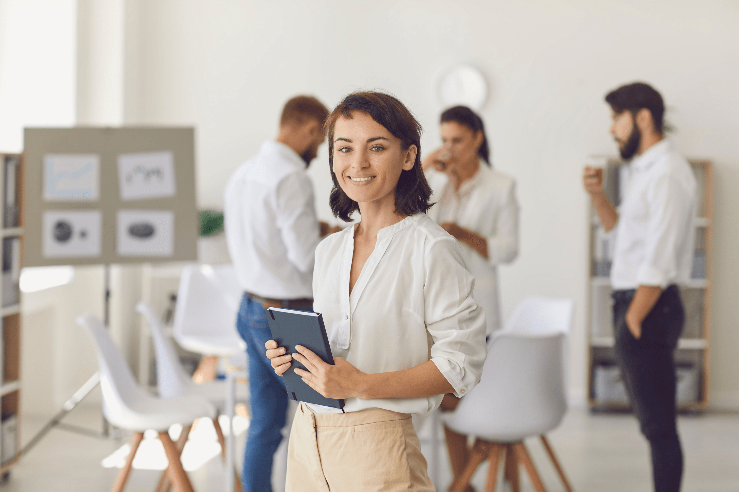 A woman with short brown hair, wearing a cream blouse and beige pants, smiling and holding a tablet, standing in an office with four people in the background.