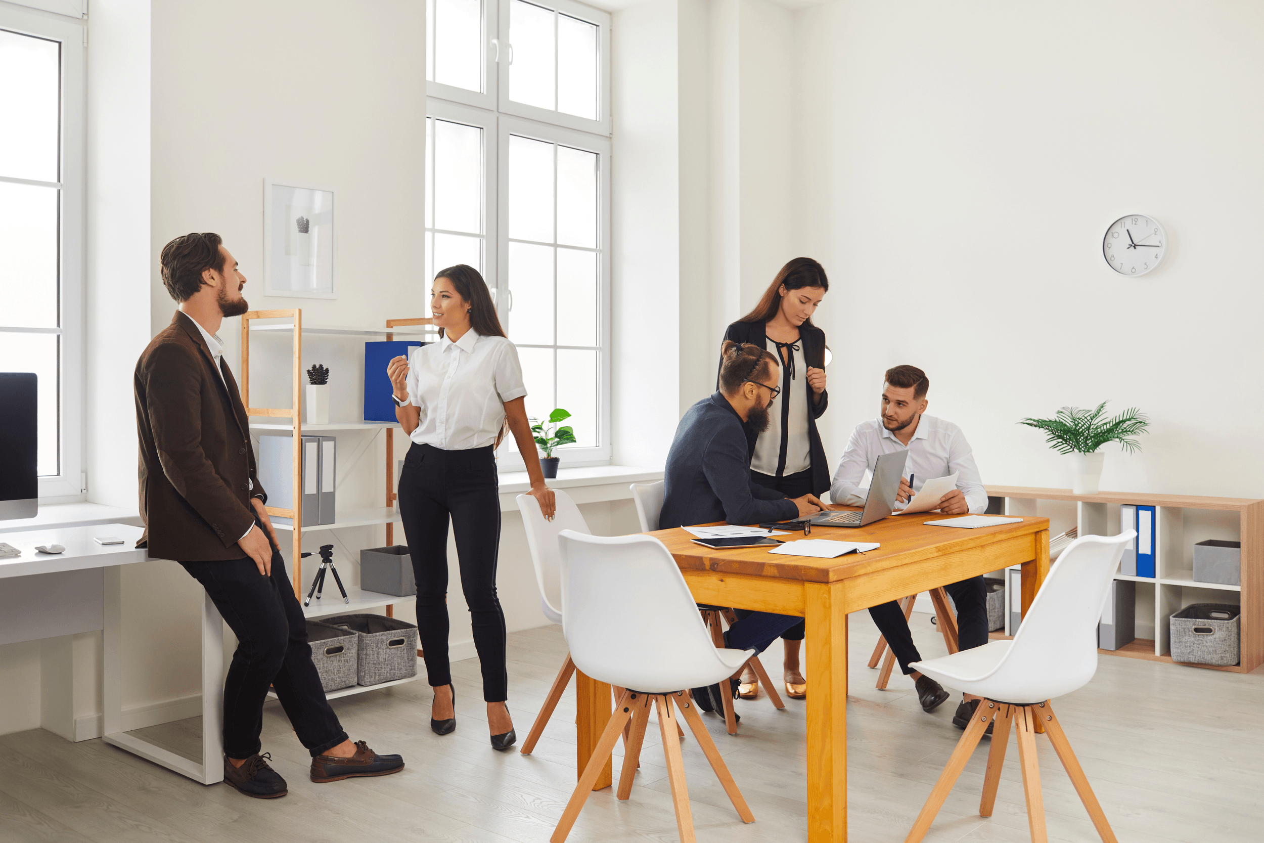 A group of professionals in an office having a meeting. Five people are engaged in conversation and work around a wooden table, with large windows letting in natural light.
