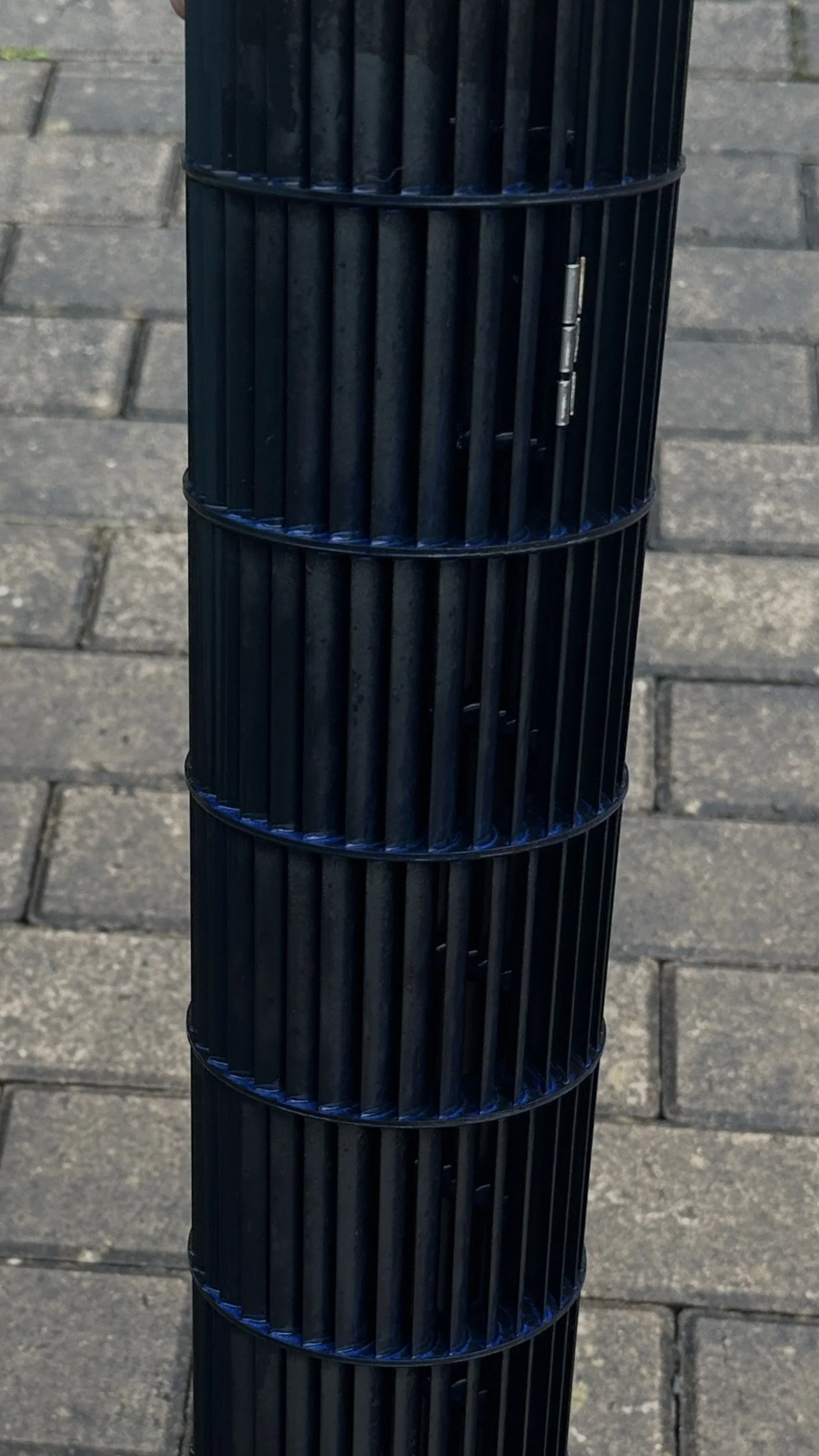 Close-up view of a black cylindrical trash can with vertical slats, mounted on a paved sidewalk.