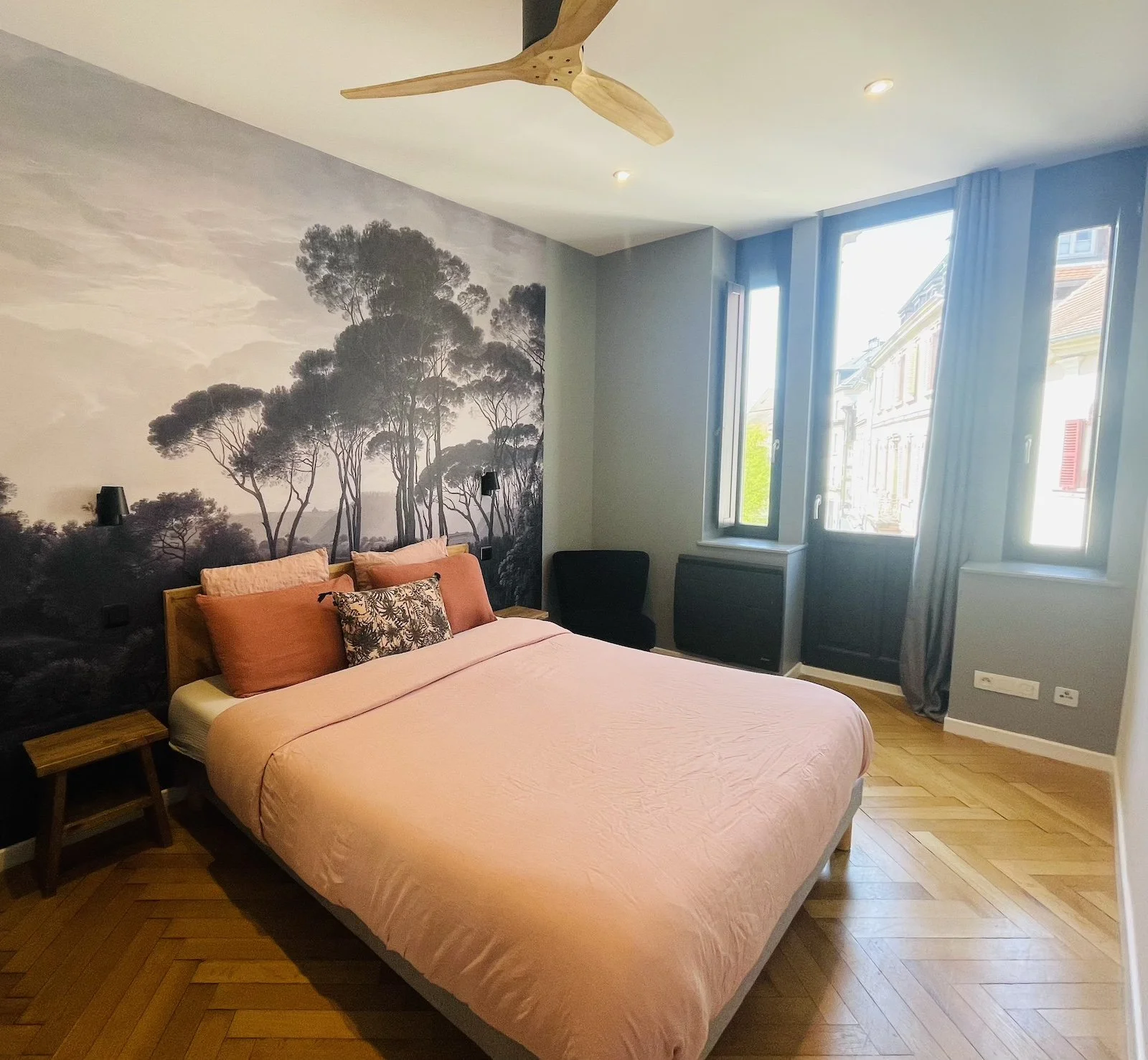 Chambre avec un lit rose, mural avec un paysage d'arbres en noir et blanc, fenêtres laissant passer la lumière, meubles en bois et un fauteuil noir au Cathedral View à Colmar en Alsace.