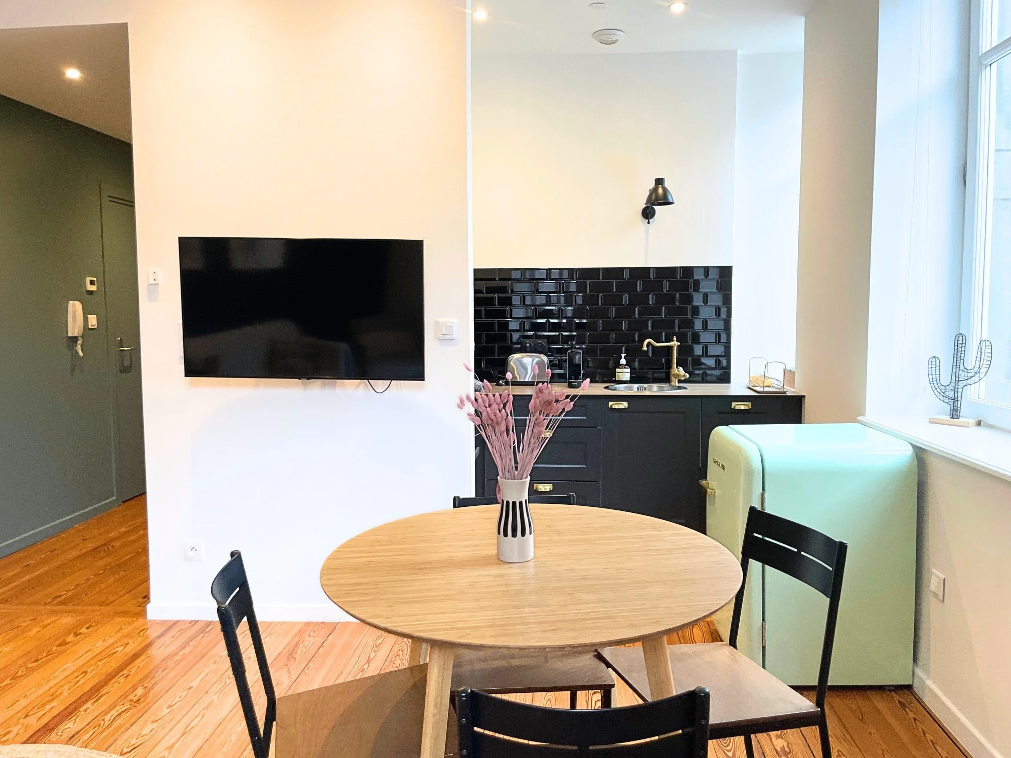 Intérieur d'une cuisine moderne avec table ronde en bois, chaises noires, téléviseur à écran plat, petite cuisine noire avec un mur en carreaux noirs, réfrigérateur vert pastel, décoration cactus en fil métallique sur le rebord de la fenêtre, et une 