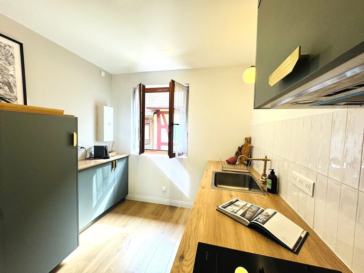 Une cuisine lumineuse à l'Adresse Duplex d'Eguisheim avec un comptoir en bois, un évier, une fenêtre ouverte laissant entrer la lumière et donnant sur les magnifiques maisons à colombages colorées des remparts.