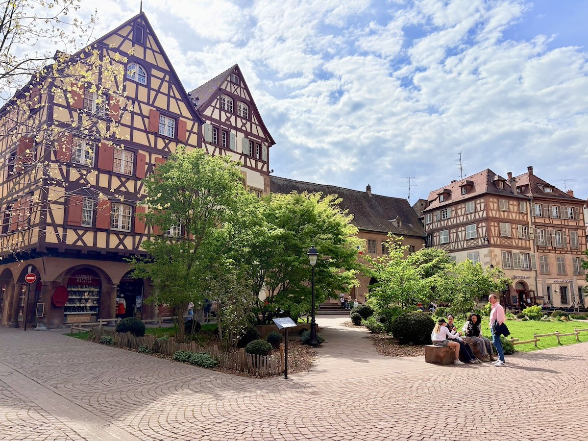 Une place pavée avec des arbres, des bancs et des personnes assises ou discutant. En arrière-plan, des bâtiments à colombages de style alsacien.