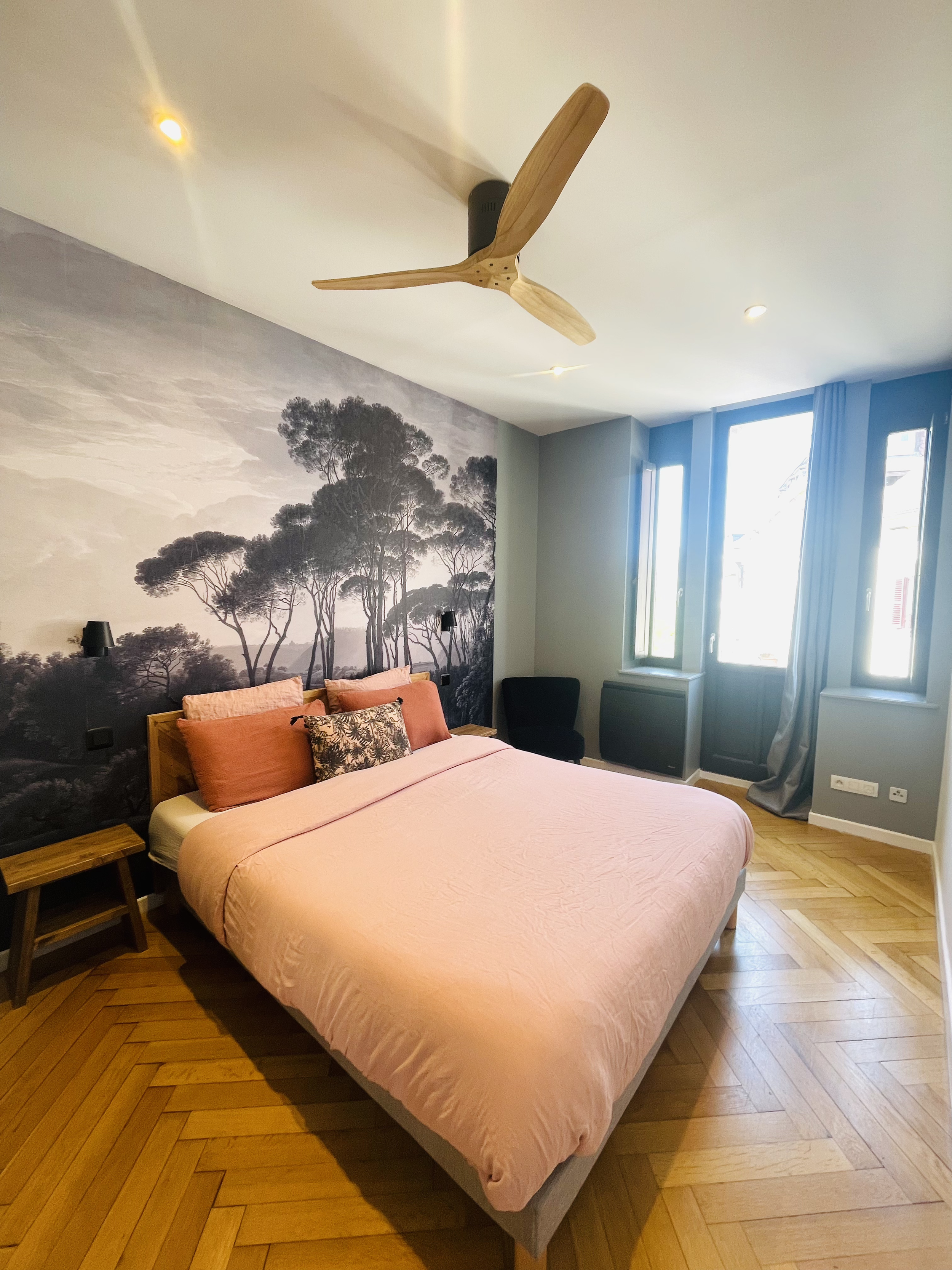 Chambre avec un lit à draps roses, un mur avec une murale d'arbres, et une fenêtre laissant entrer la lumière