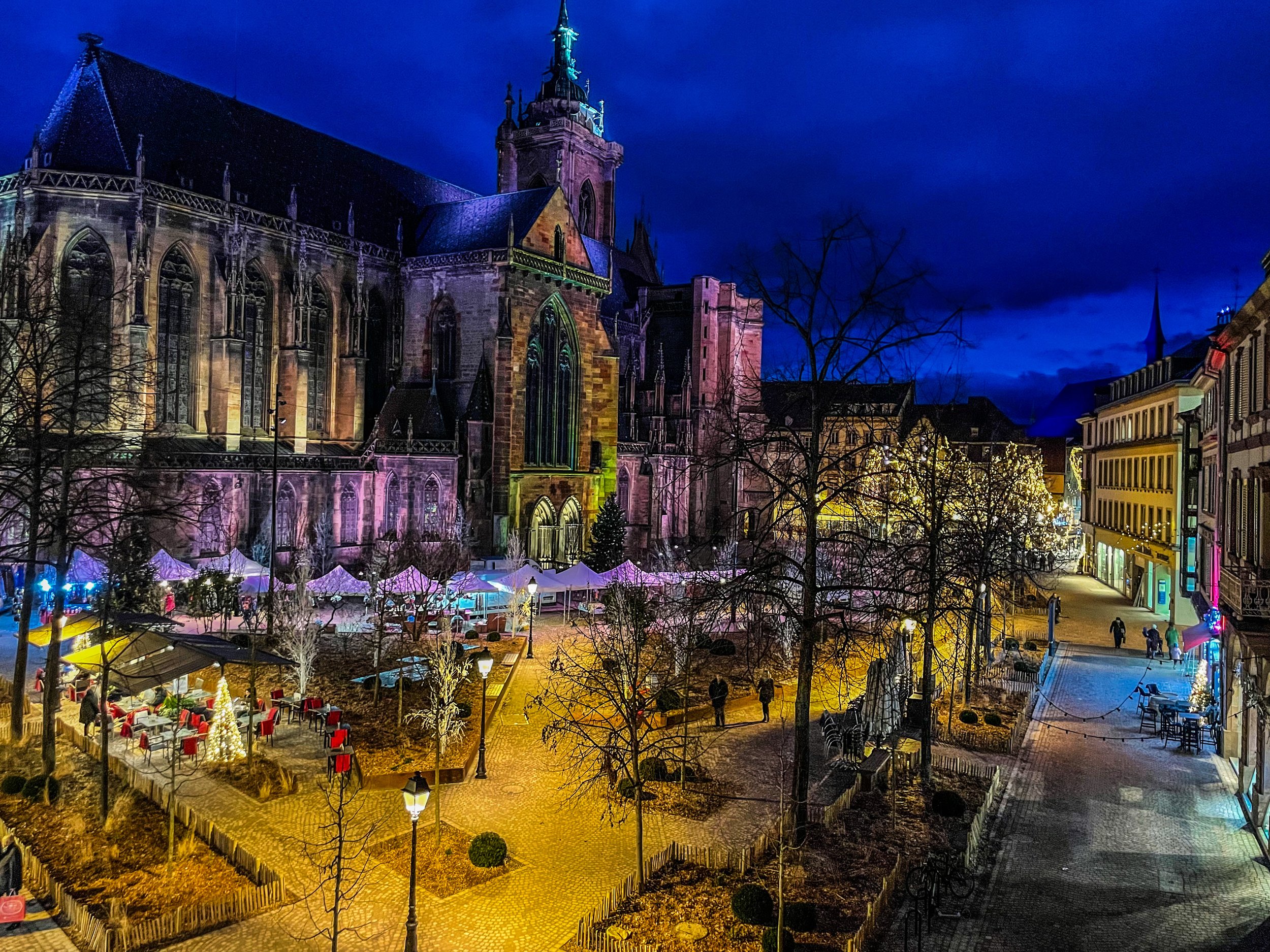 Vue nocturne d'une place de ville avec une grande église gothique illuminée, des arbres sans feuilles, des lampadaires, des tables de marché de Noël avec des tentes blanches, et des bâtiments colorés bordant la rue.
