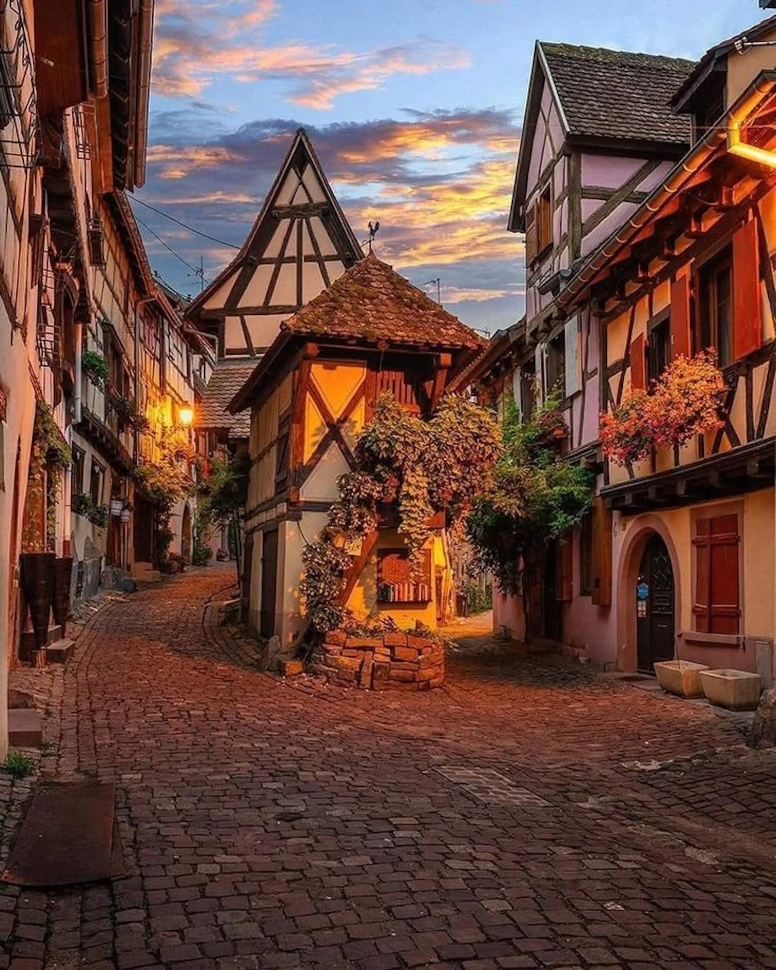 Ruelle pavée avec des maisons à colombages, fleurs et arbre en fleurs au crépuscule, ambiance chaleureuse.