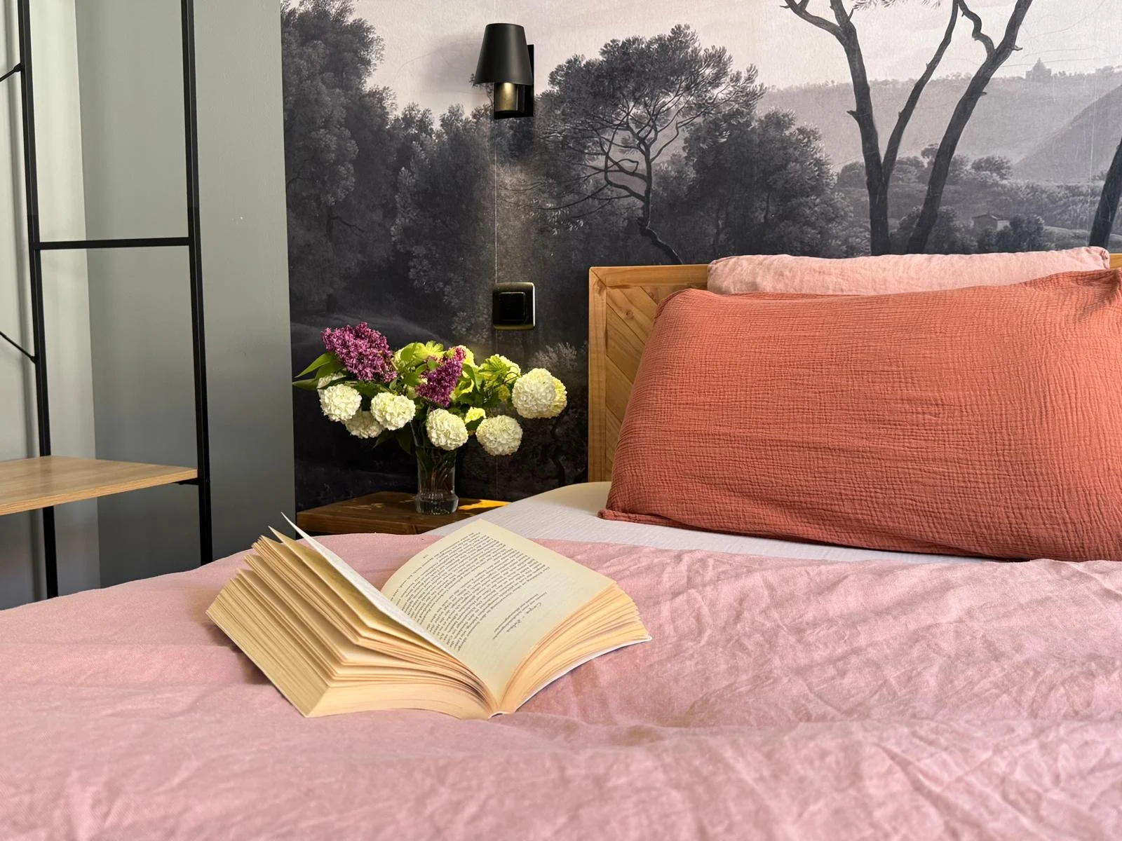 Un lit avec des draps rose et des oreillers, une table de chevet avec un vase de fleurs violettes et blanches, une lampe noire, un mur en arrière-plan avec une peinture de paysage forestier en noir et blanc au Cathedral View à Colmar.