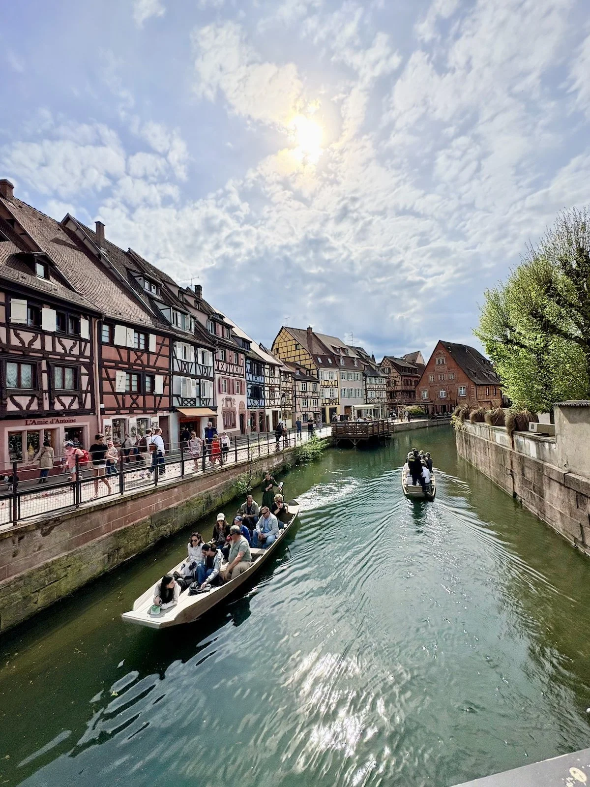 Vue d'une rue avec des maisons à colombages colorées le long d'une rivière, où passent deux bateaux avec des passagers par une journée ensoleillée.