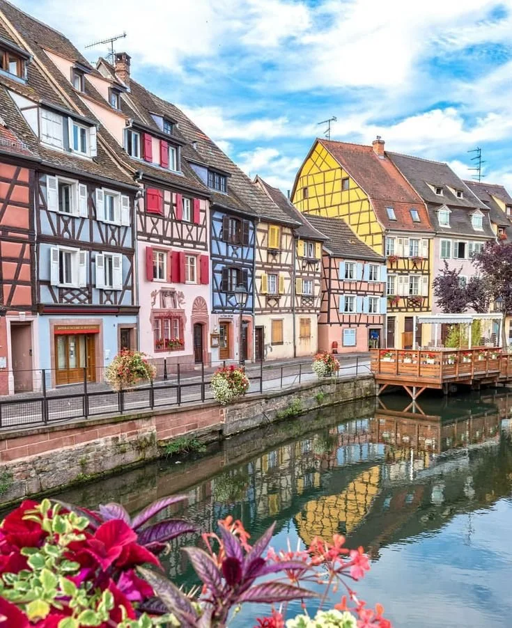 Maisons colorées à colombages le long d'une rivière, avec un ciel bleu, fleurs au premier plan et un pont en bois.