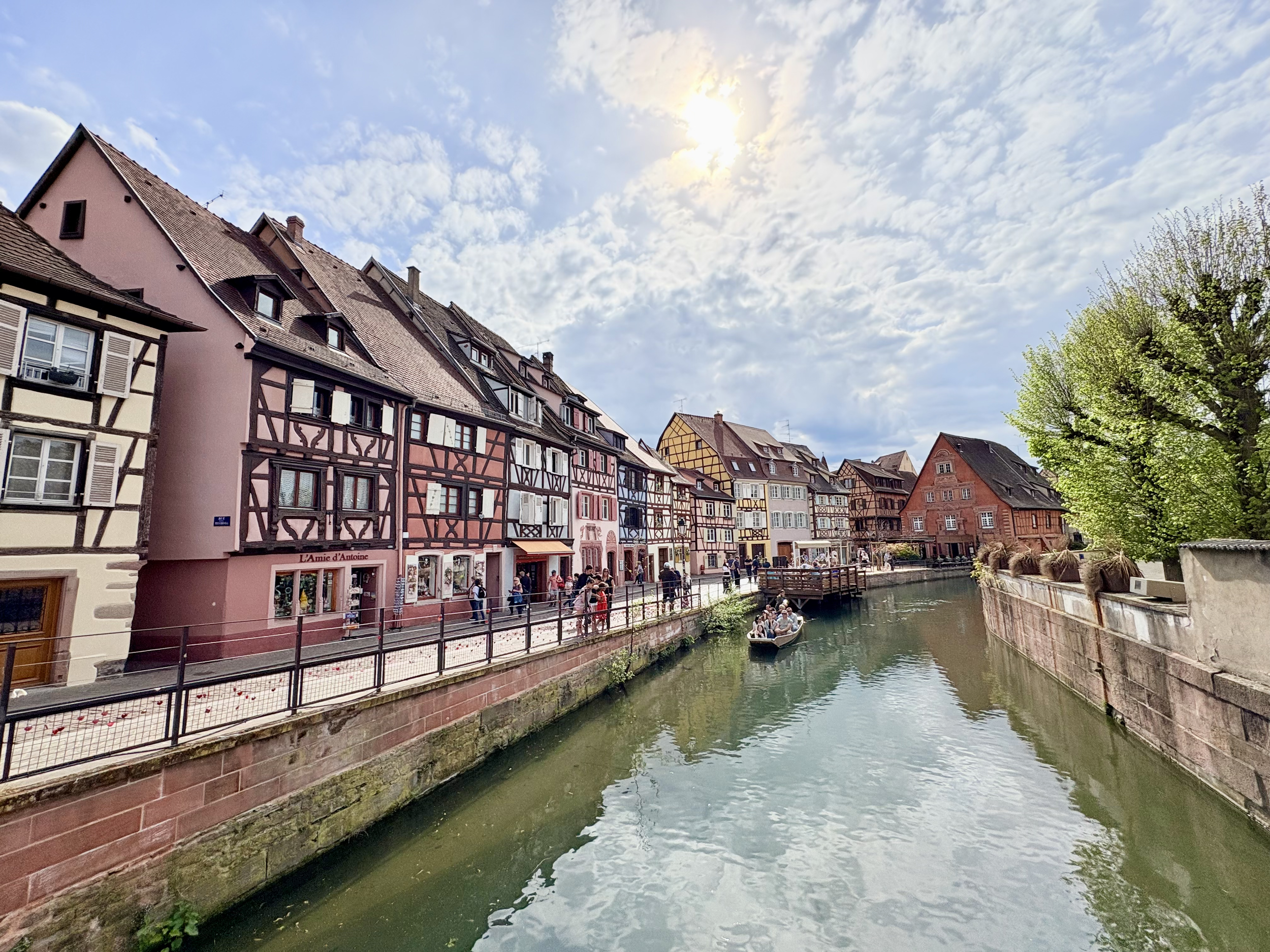 Vue panoramique d'une ville avec des maisons à colombages colorées le long d'une rivière, avec un bateau naviguant et un ciel partiellement nuageux sur la Petite Venise de Colmar proche du Cathedral View.
