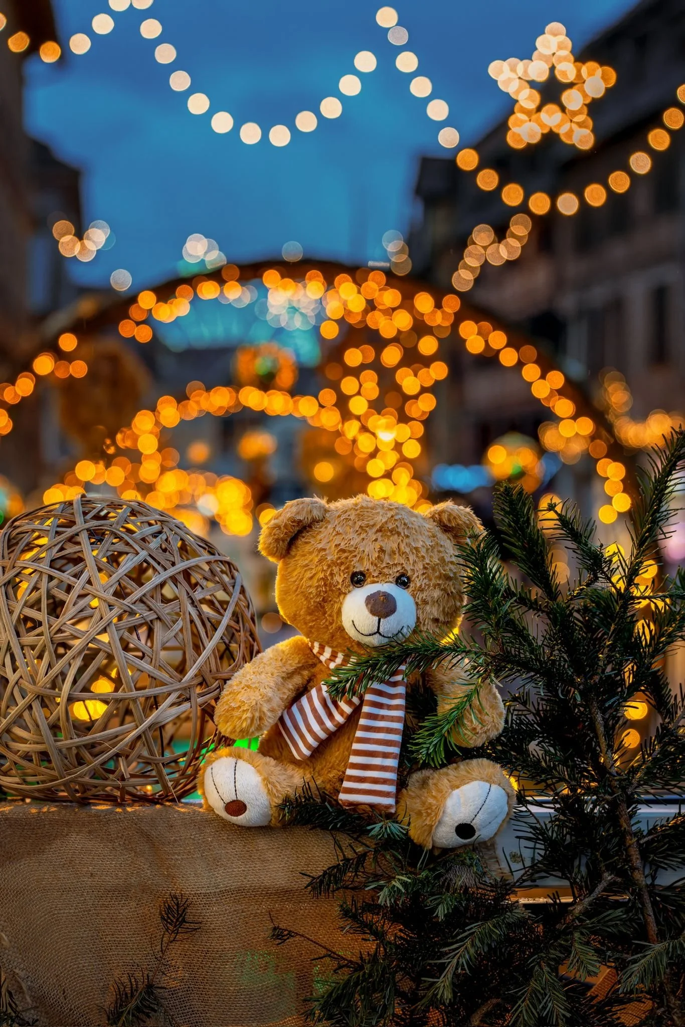 Un ours en peluche beige avec un foulard à rayures brune et blanche, une branche de sapin, et un objet décoratif en forme de sphère en rotin, tous placés sur une surface en tissu, avec un décor de fêtes en arrière-plan éclairé de guirlandes lumineuse