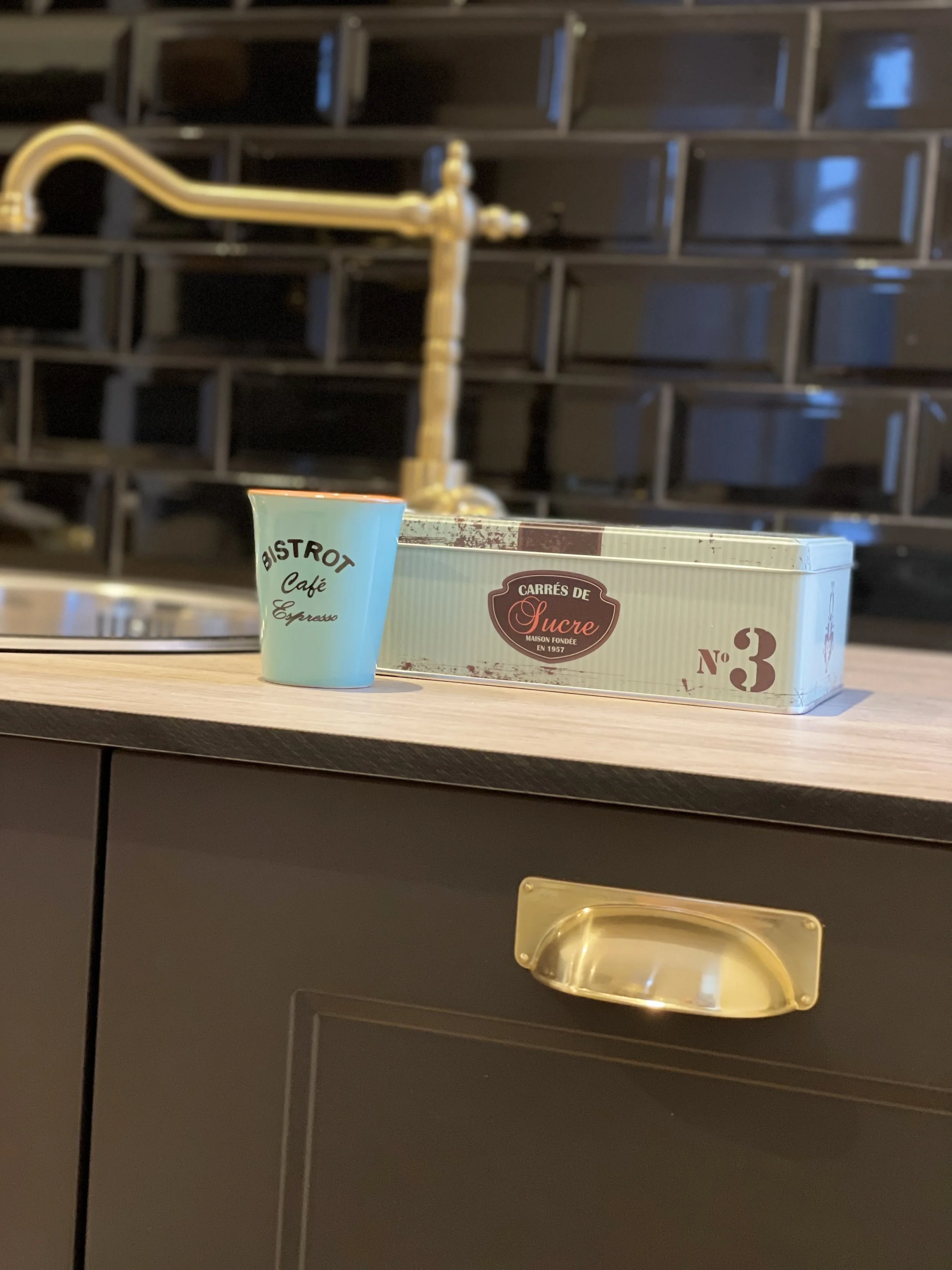 Une petite tasse en papier bleu avec l'inscription 'BISTROT Café Espresso' à côté d'une boîte en carton vintage sur un comptoir en bois. En arrière-plan, un robinet de cuisine en laiton et un mur en carreaux noirs.