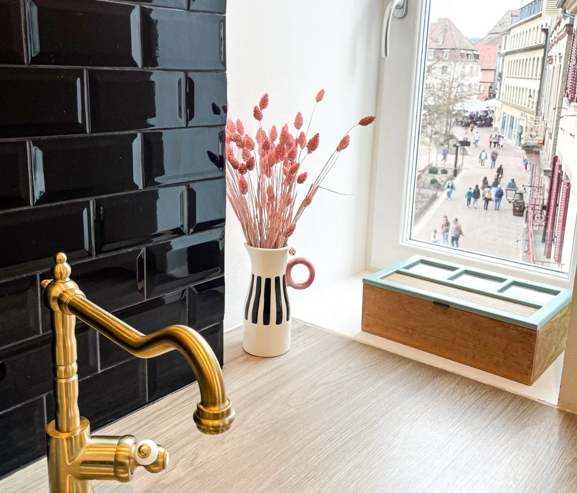 Intérieur avec un vase de fleurs séchées roses, une fenêtre donnant sur une rue piétonne, un meuble en bois sous la fenêtre, et un mur noir brillant en carrelage, avec un robinet en laiton visible.