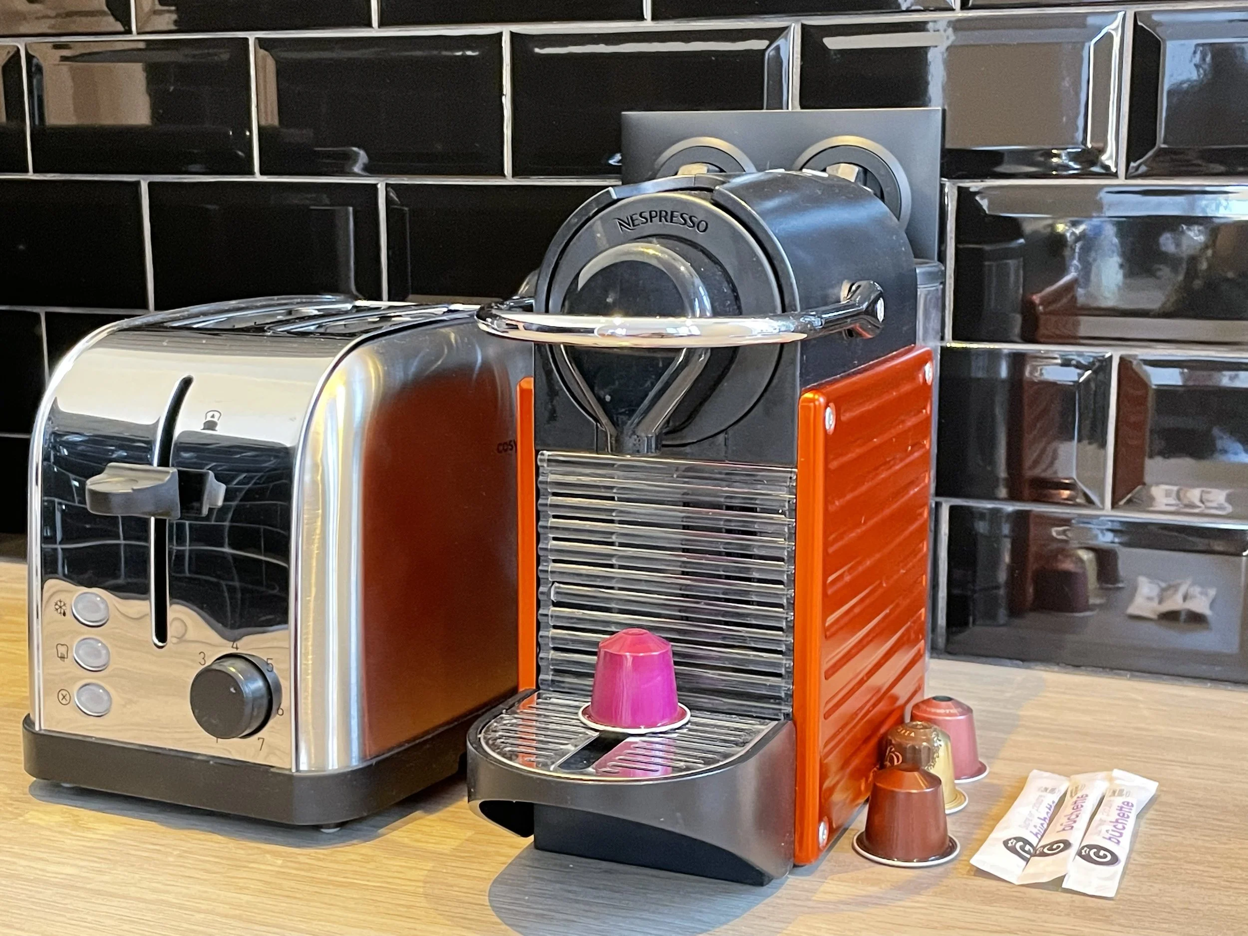 Une machine à café Nespresso avec un support pour capsules et une grille de récupération, accompagnée de plusieurs capsules de café de différentes couleurs, sur une table en bois.
