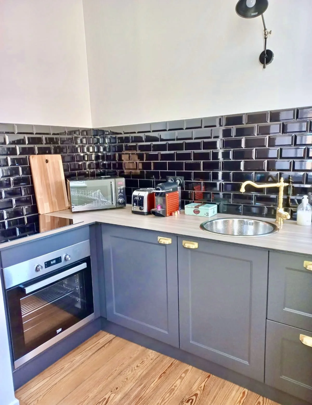 Coin cuisine moderne avec four, micro-ondes, grille-pain, kettles, plan de travail en bois, évier en métal, dosseret en carrelage noir brillant, porte-voiture en bois, lumière suspendue noire, bouteille de savon.