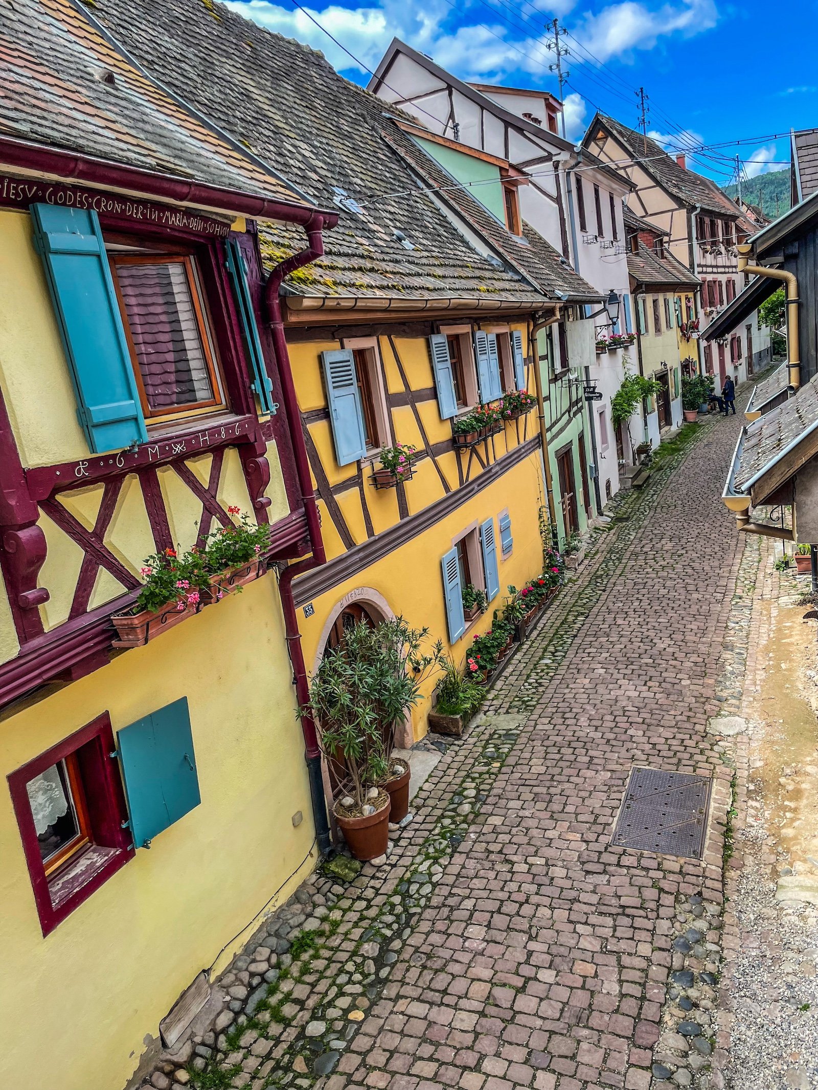Rue pavée bordée de maisons colorées avec volets bleus et fleurs suspendues, ciel bleu avec quelques nuages.