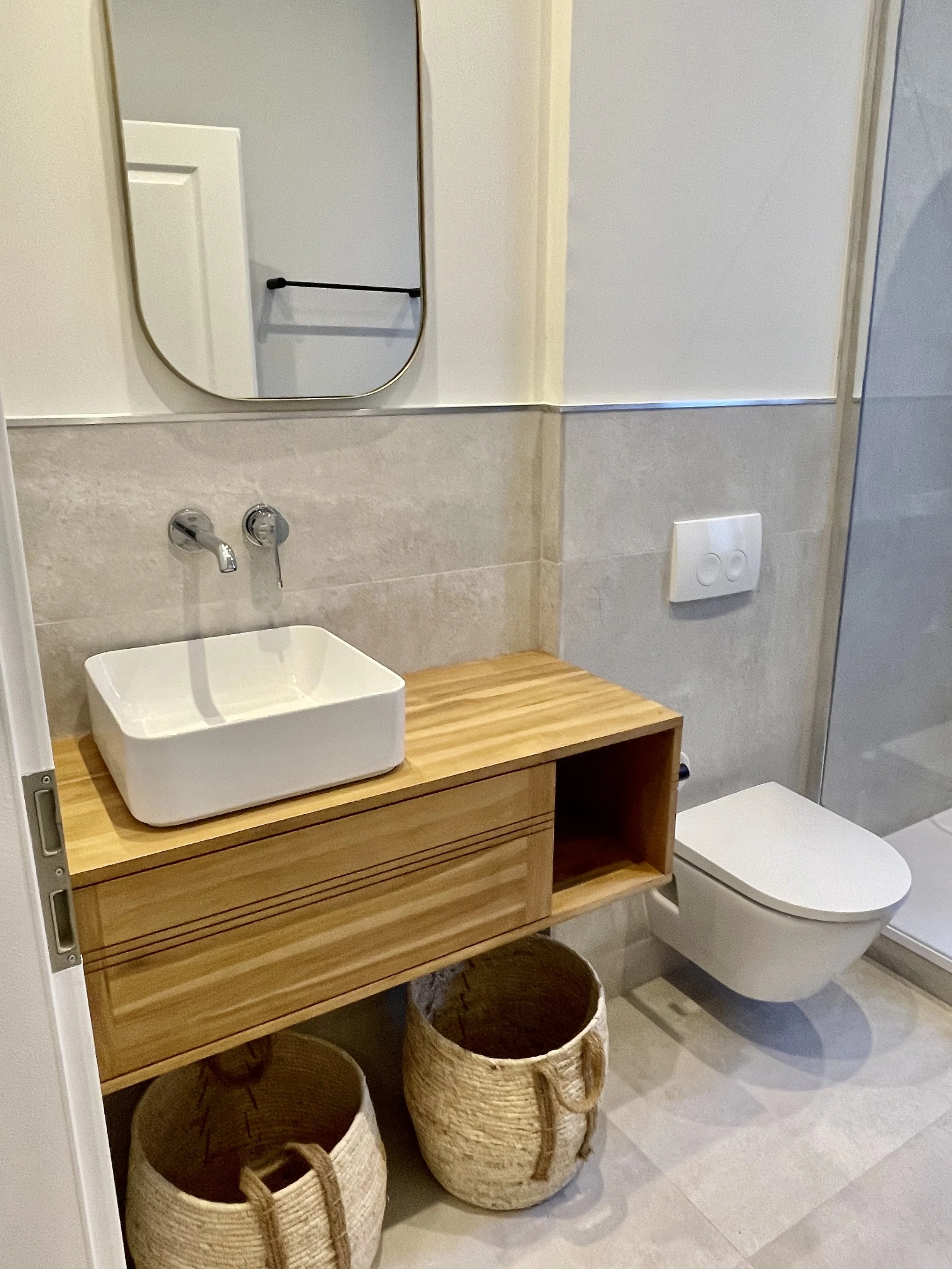 Petite salle de bain avec un lavabo en forme de caisse blanche placé sur une console en bois, un miroir rectangulaire au-dessus, un toilette suspendu à droite, un évier en dessous du miroir, et deux paniers en osier sous la console.