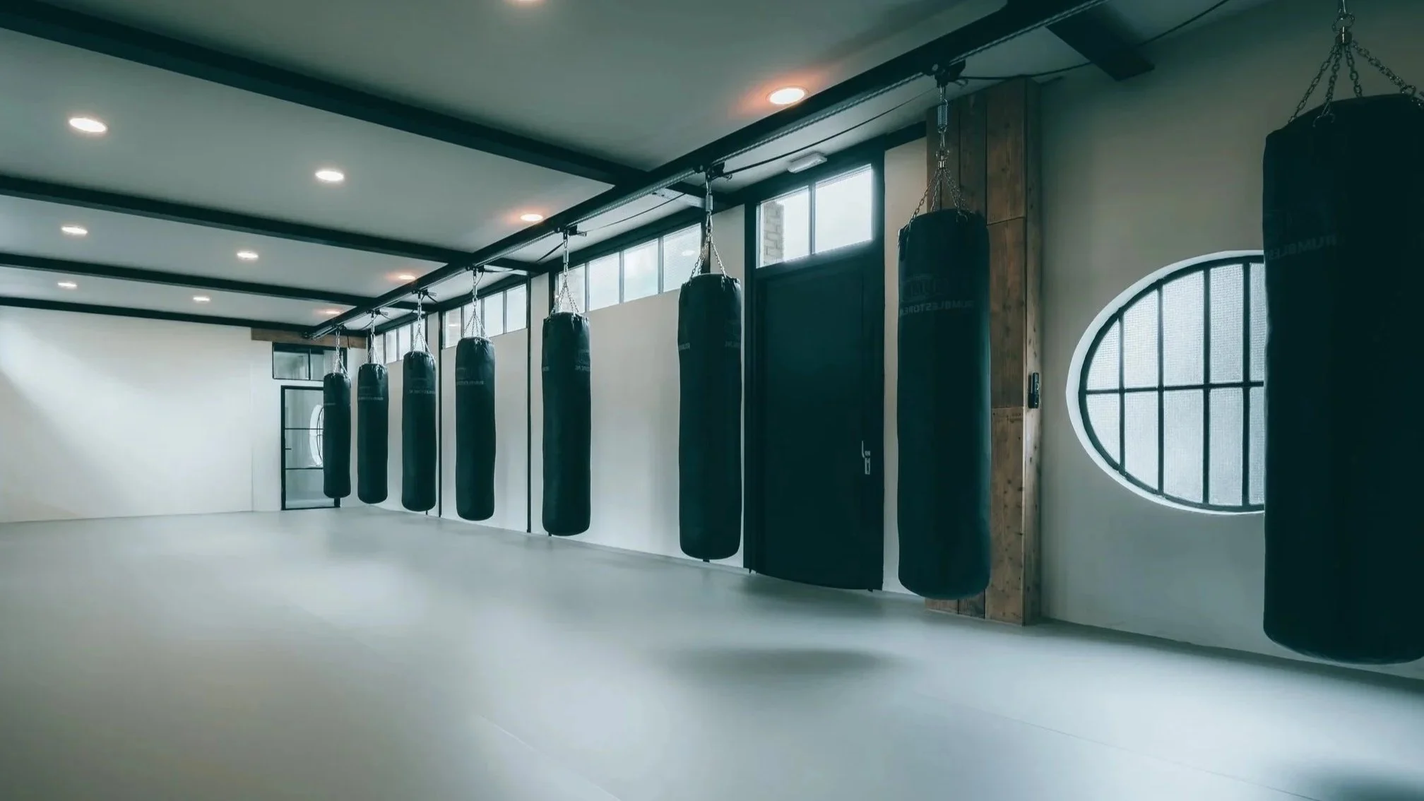 Binnenruimte van een fitnessruimte met zwart hanging bokszakken, grote ramen en een witte vloer.