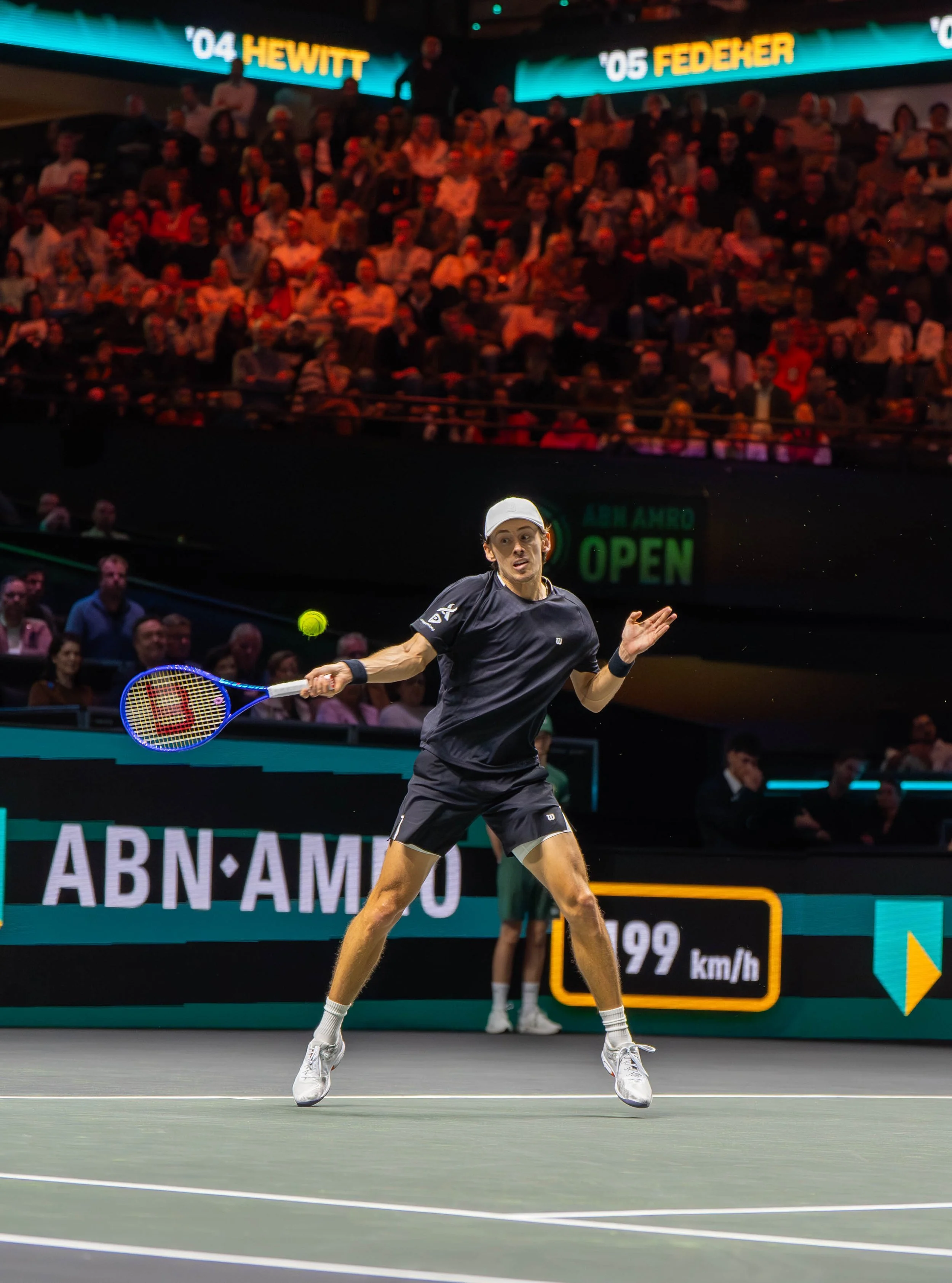 A male tennis player in a black outfit and white cap hits a backhand shot during a match in an indoor stadium with a crowd of spectators watching.