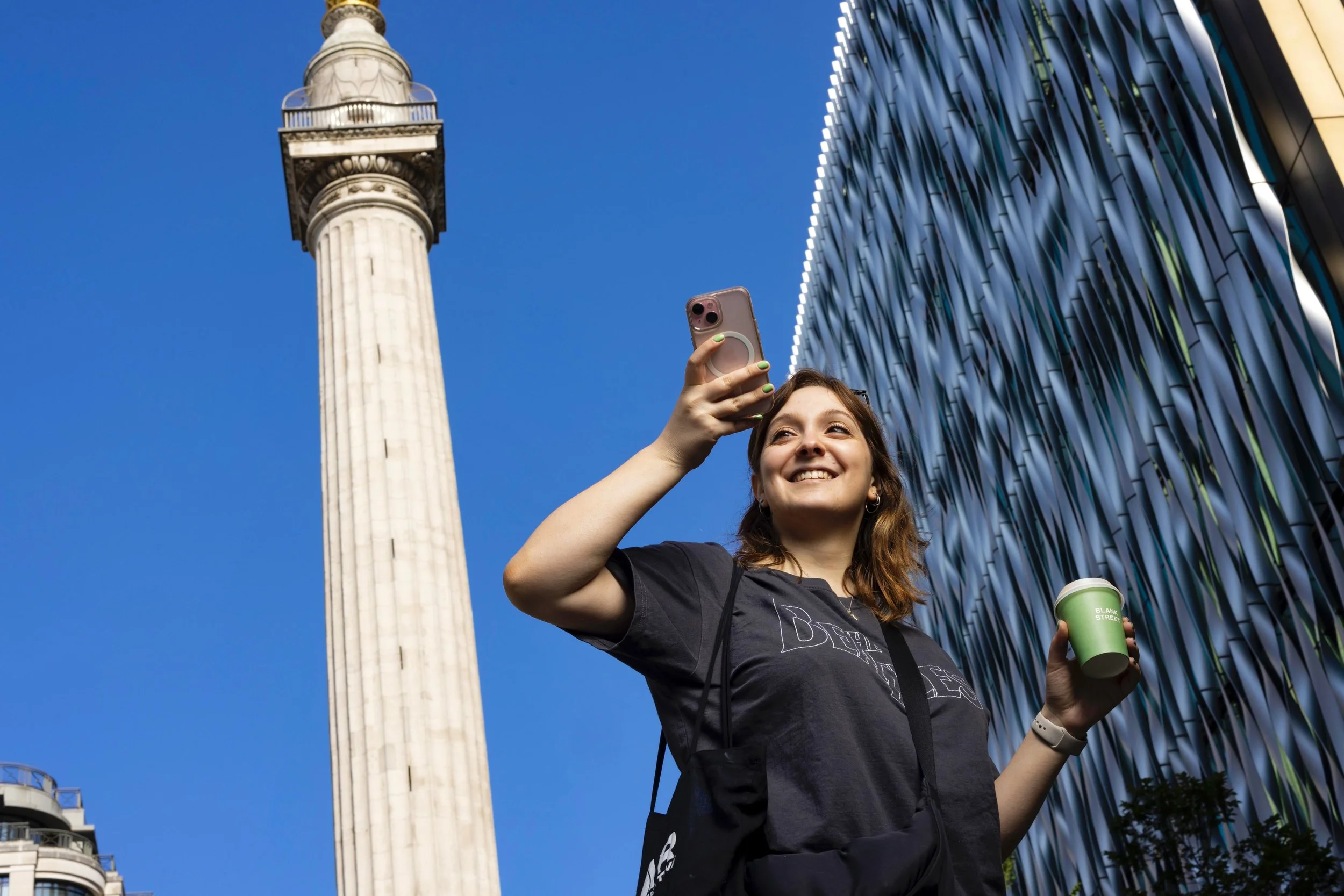 Young woman taking a selfie with a smartphone on a city street, holding a green coffee cup, with the Washington Monument and modern building in the background.