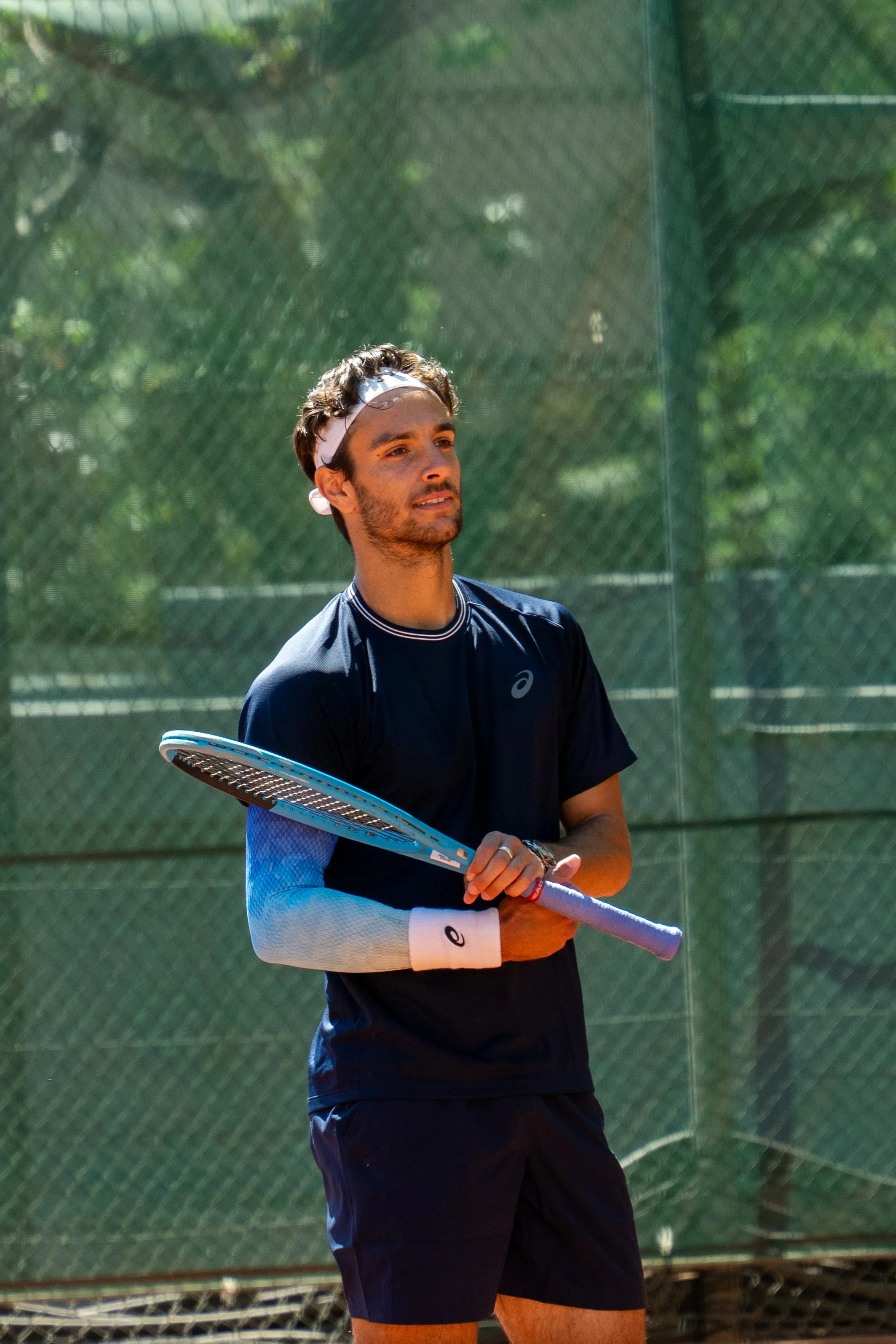 Lorenzo Musetti, wearing a dark sports shirt with elbow sleeves and a white headband, holds a tennis racquet on an outdoor court, with a green chain-link fence and trees in the background.