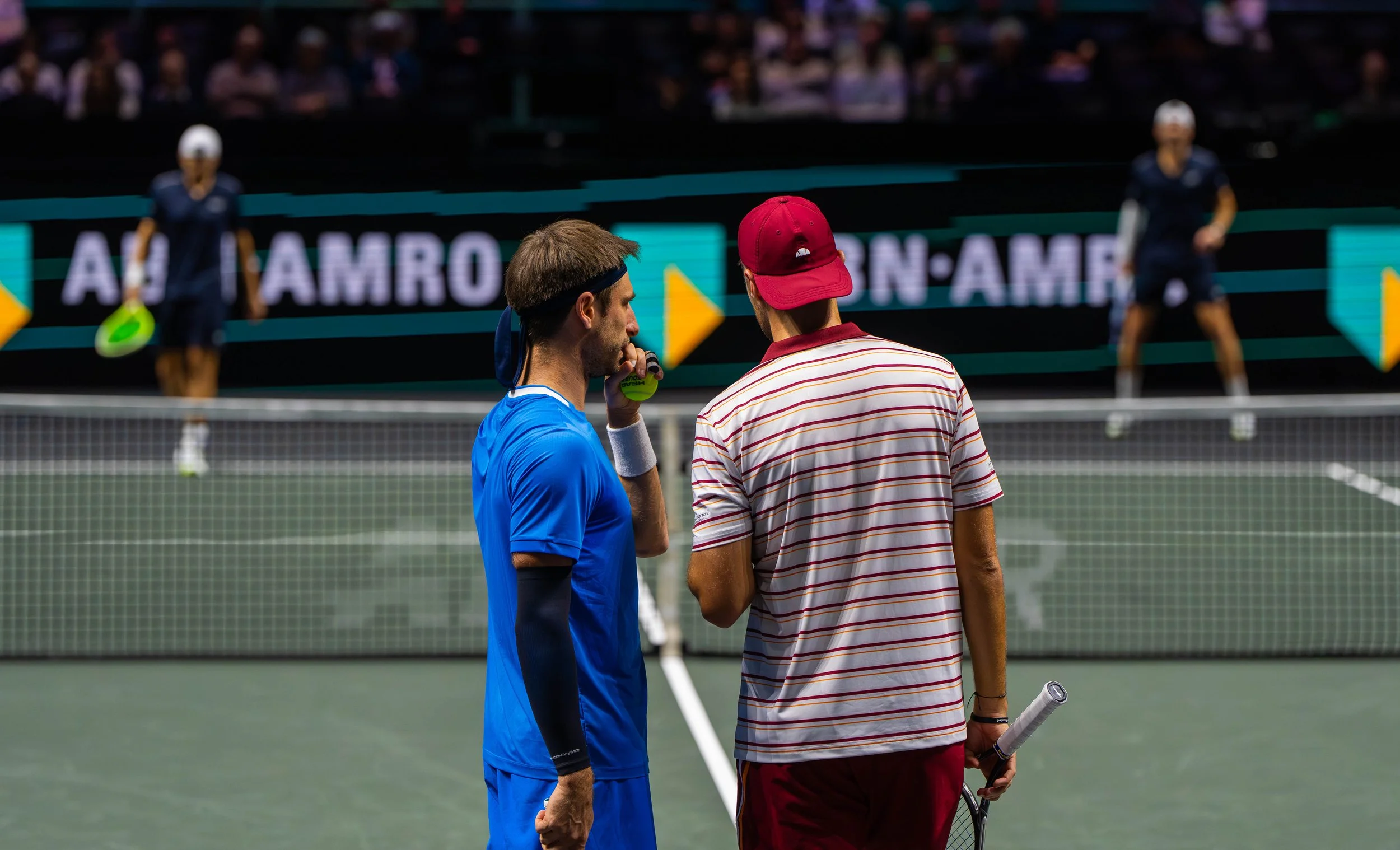 Two male tennis players standing on a tennis court, engaged in conversation, with two female players in the background, one holding a tennis racket, during a match or practice session.