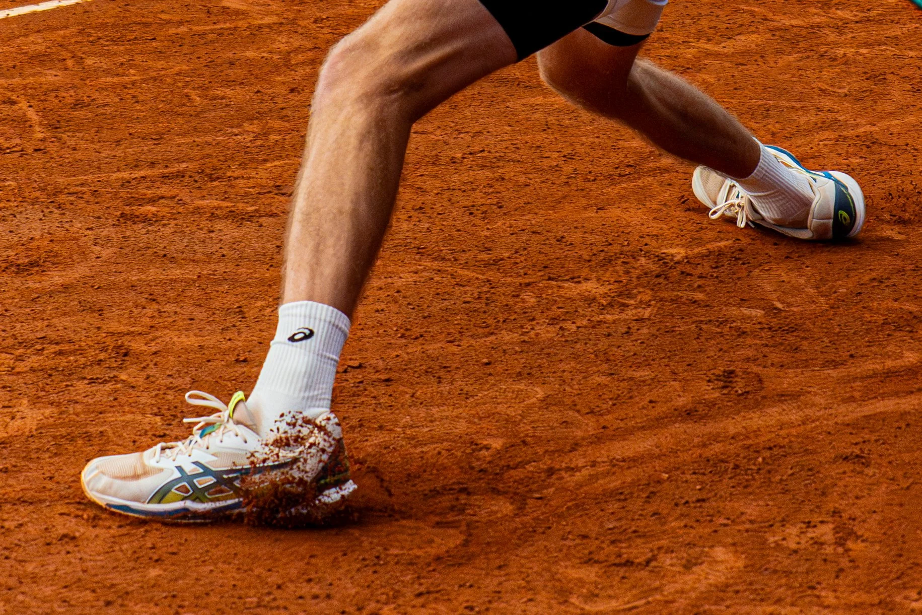 Close-up of a tennis player's legs and shoes on a clay court, with the player lunging forward and kicking up dirt.
