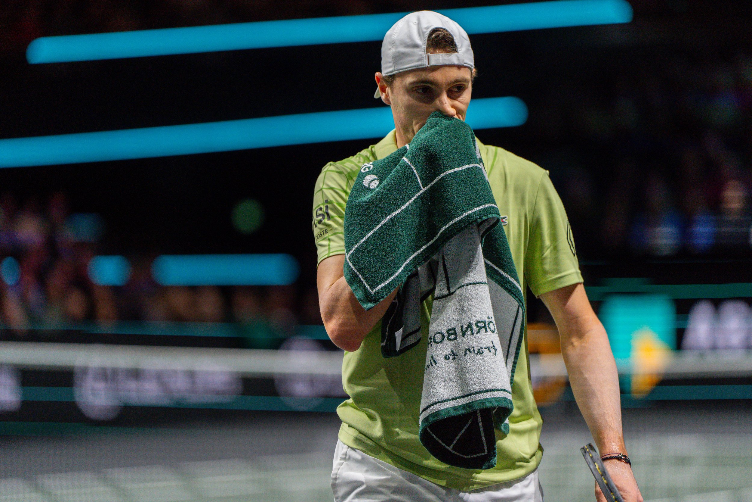 A male tennis player holding a towel to his face on an indoor hard court during a match.