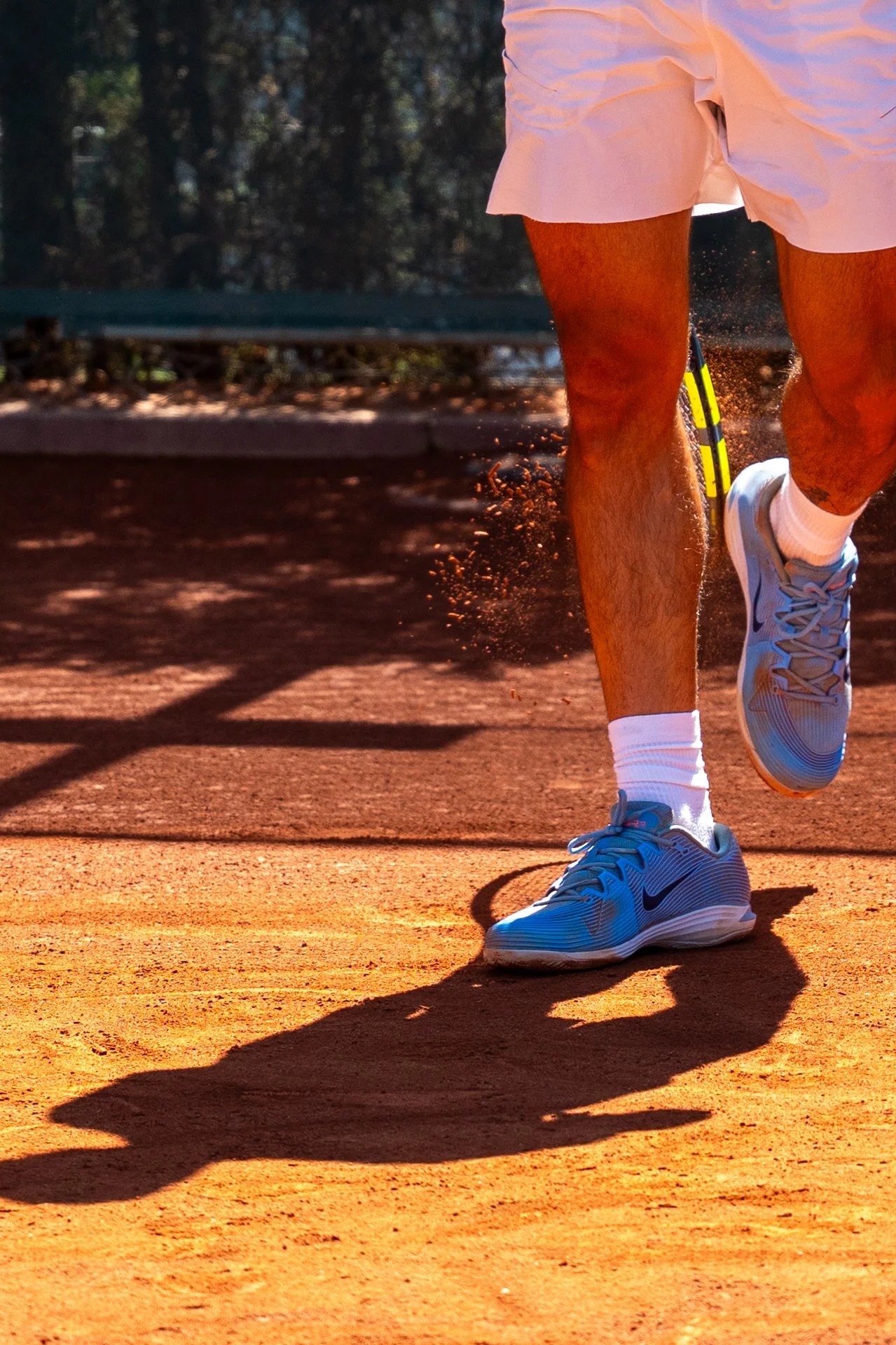 Side view of Carlos Alcaraz wearing white sneakers, white socks, white shorts, and a white shirt, running on a clay tennis court during daytime, with shadows cast on the ground and a blurred background of trees.
