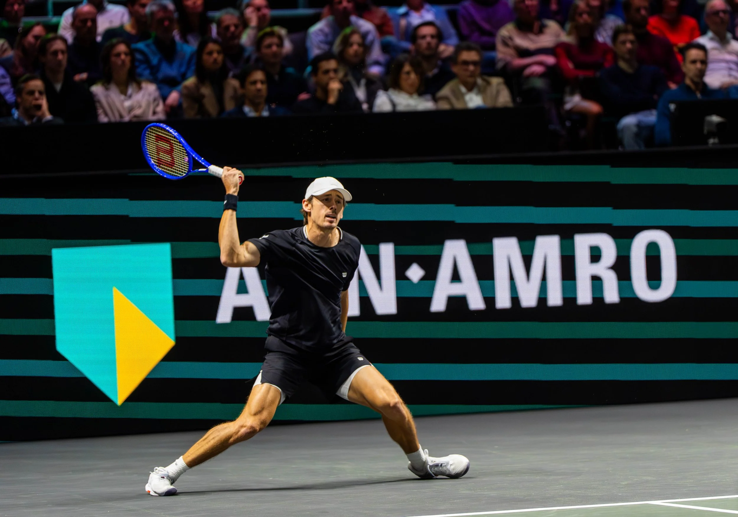 A male tennis player in black athletic wear and a white cap prepares to hit a backhand shot during a match, with an audience watching in the background and an advertisement display on the court.