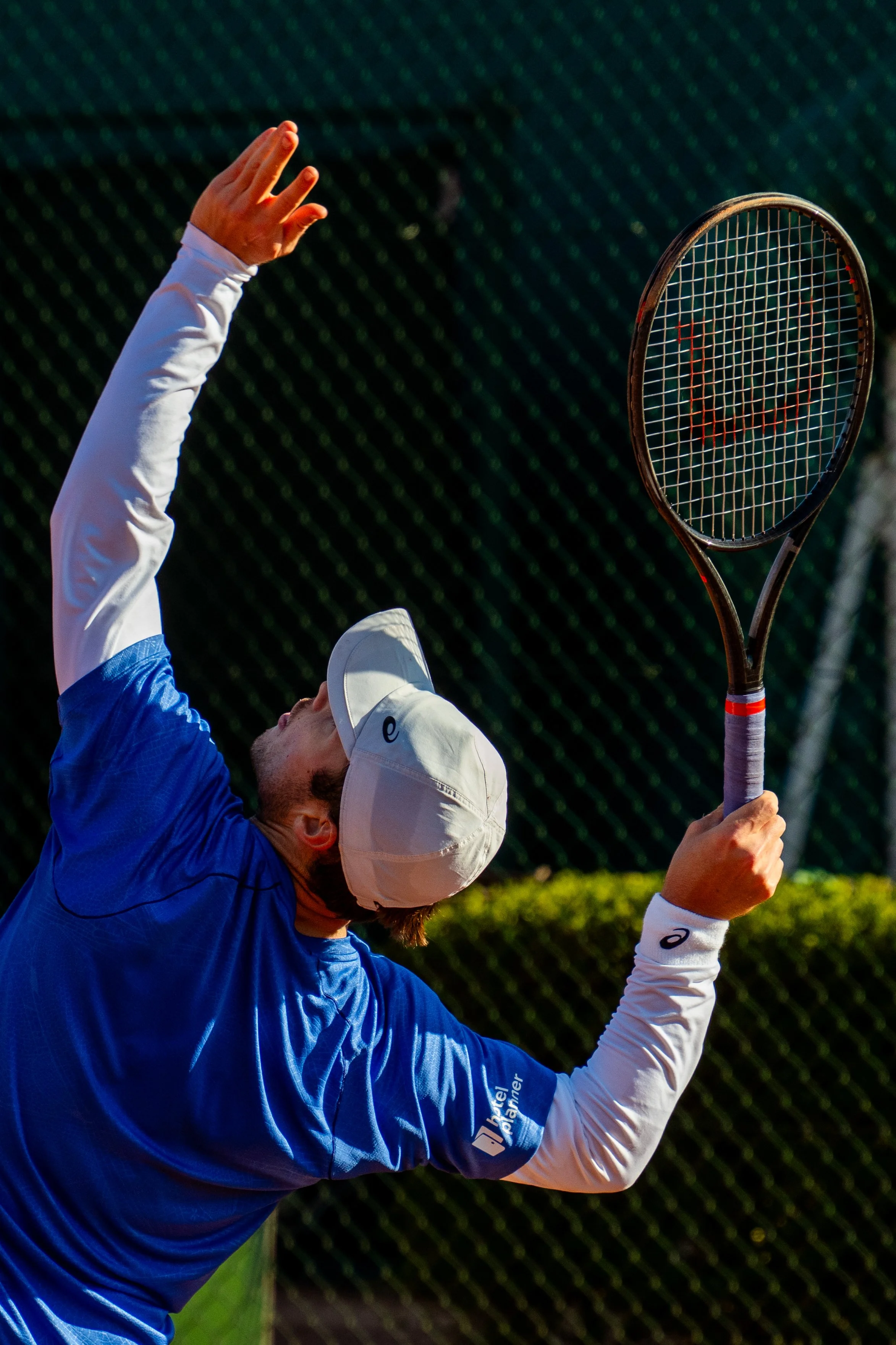 A tennis player in a blue shirt and white cap serving the ball on a tennis court.