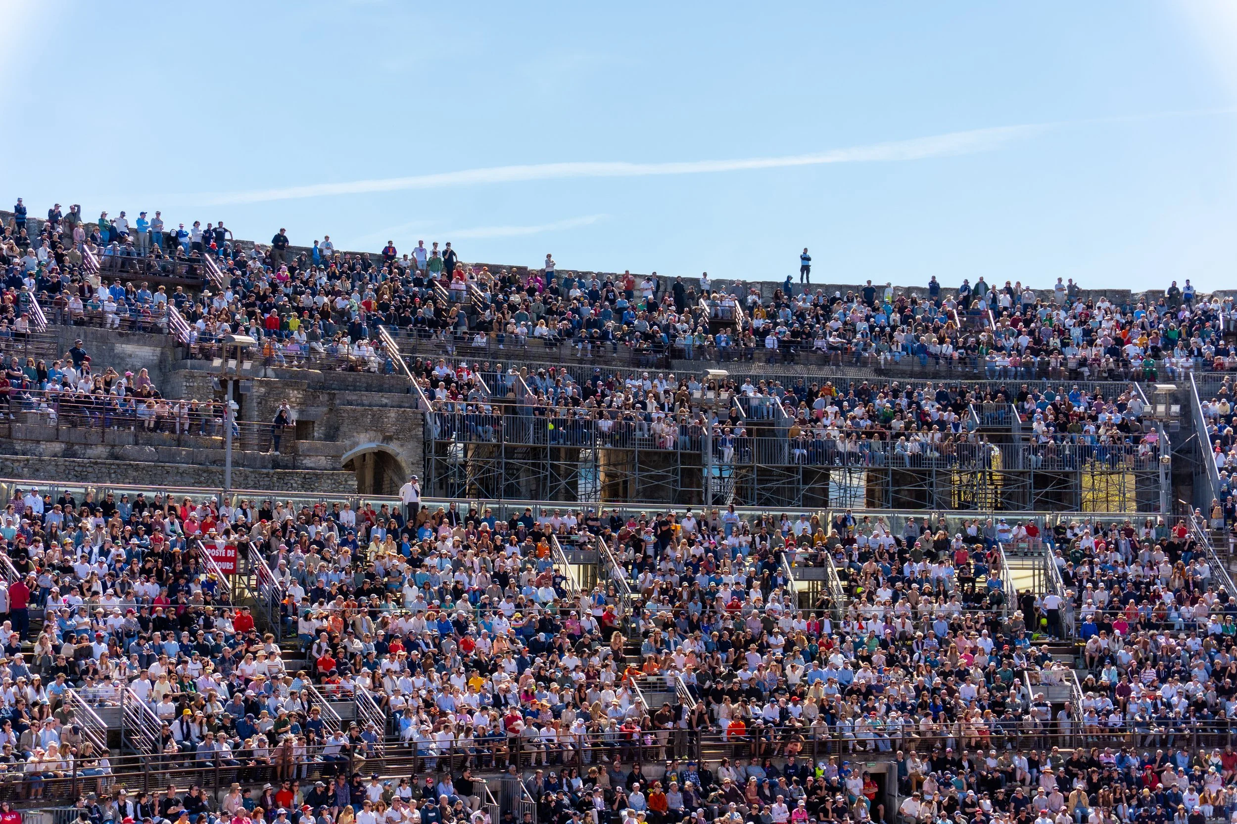 Large crowd of people sitting in stadium bleachers under a clear blue sky.