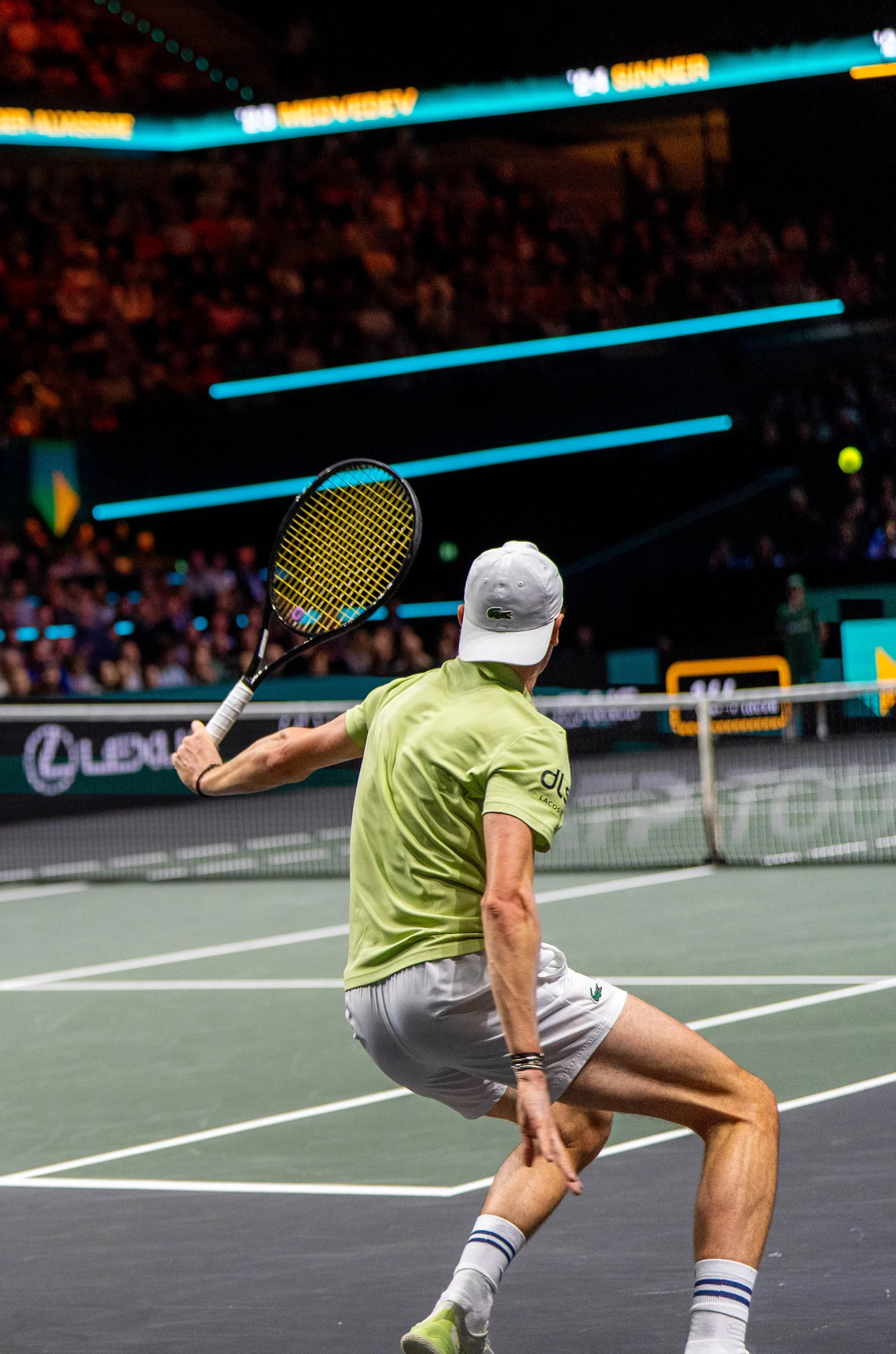 A male tennis player in a lime green shirt, white shorts, gray cap, and white socks with blue stripes, prepares to hit a tennis ball on a court with spectators and digital screens in the background.