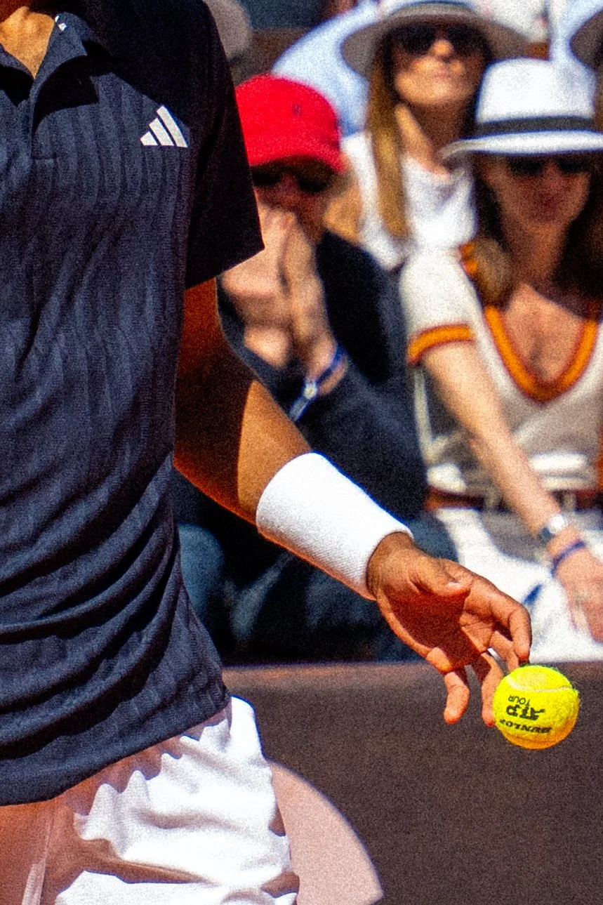 Person holding a yellow tennis ball on a tennis court, with spectators wearing sun hats sitting in the background.
