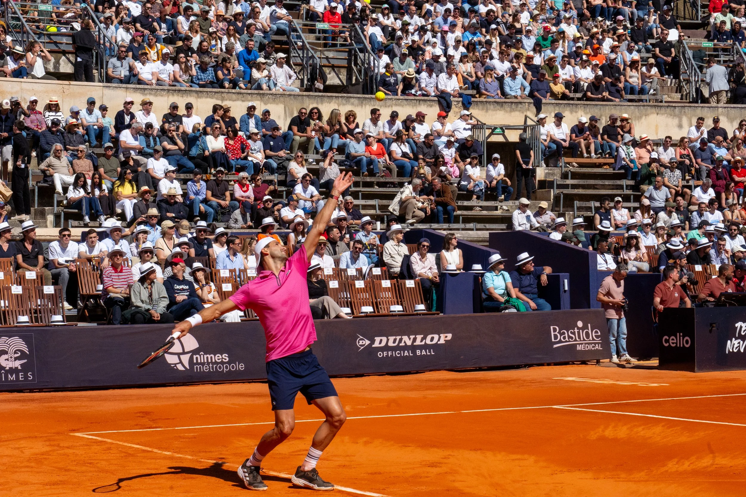 A tennis player in a pink shirt and navy shorts preparing to hit a tennis ball on a clay court, with spectators watching from the stands.