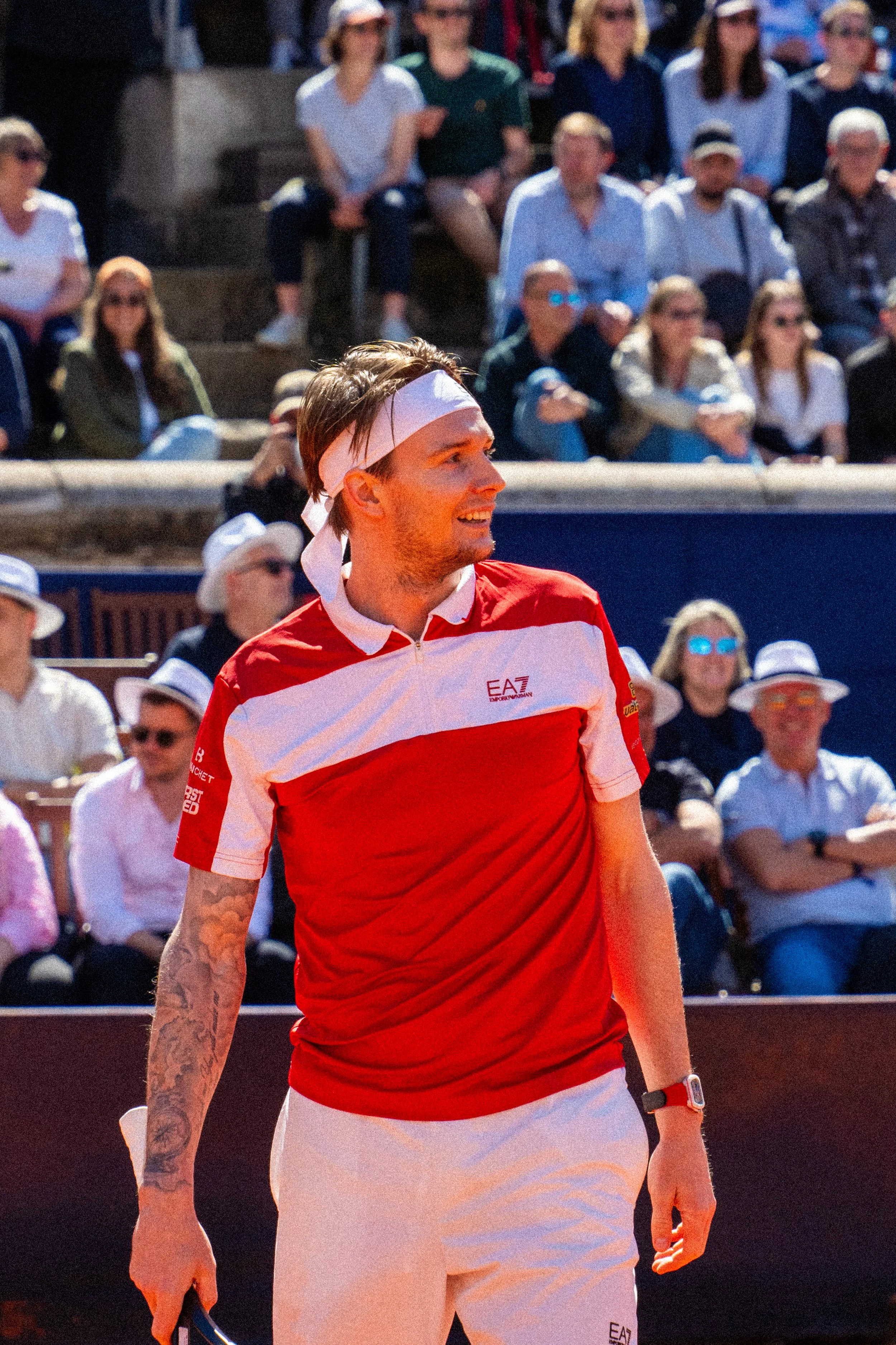 A male tennis player in a red and white shirt and white shorts, with a bandana on his head, standing on a tennis court during a match. There are spectators seated in the background watching.