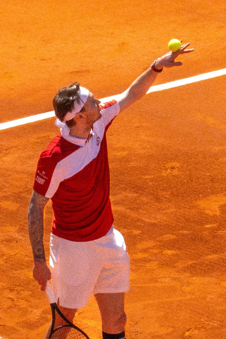 A tennis player on a clay court preparing to serve, wearing a white headband, red and white shirt, and white shorts.