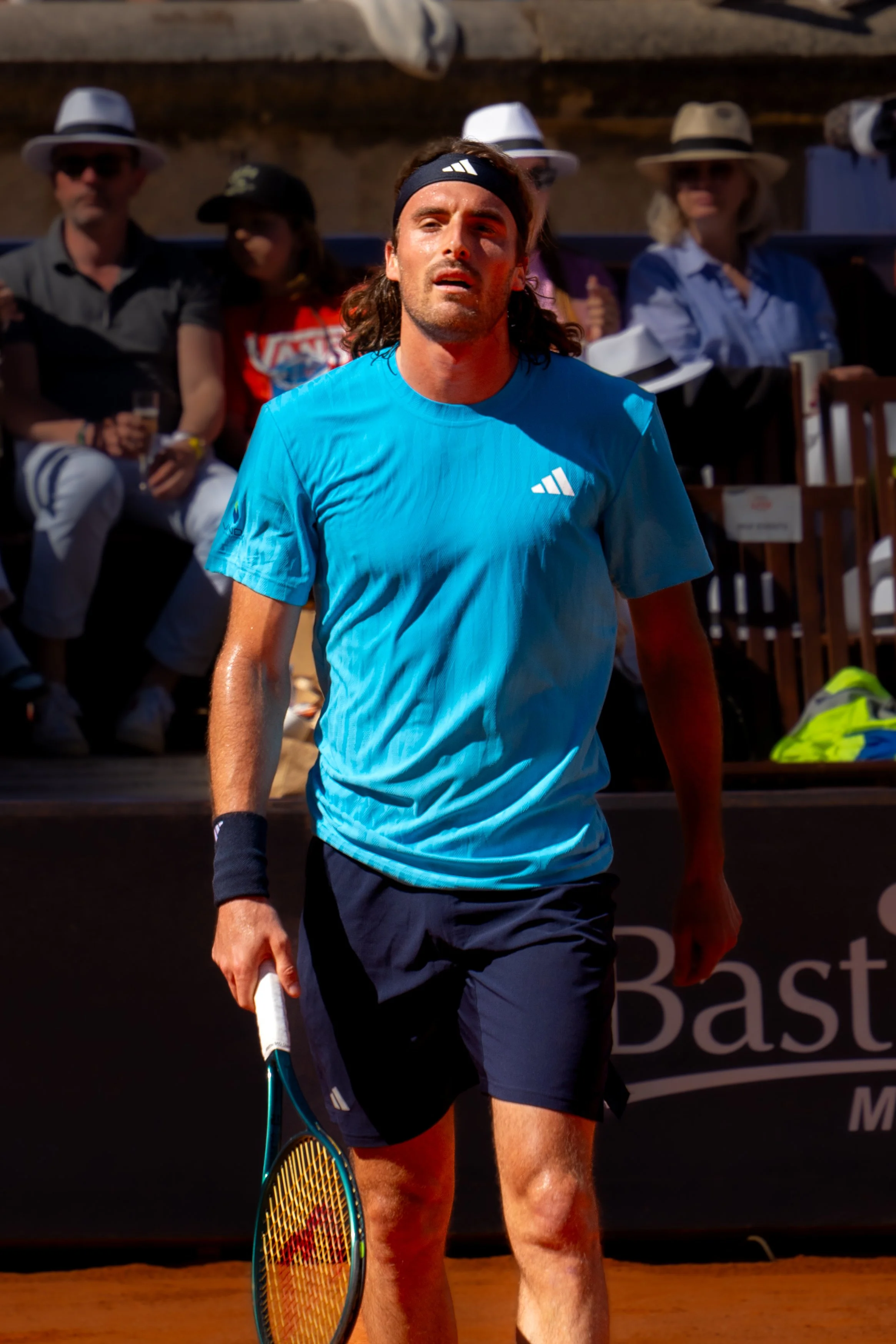 A male tennis player with long hair, wearing a blue Adidas shirt, black shorts, and a black wristband, holding a tennis racket and walking on a clay court during a match.