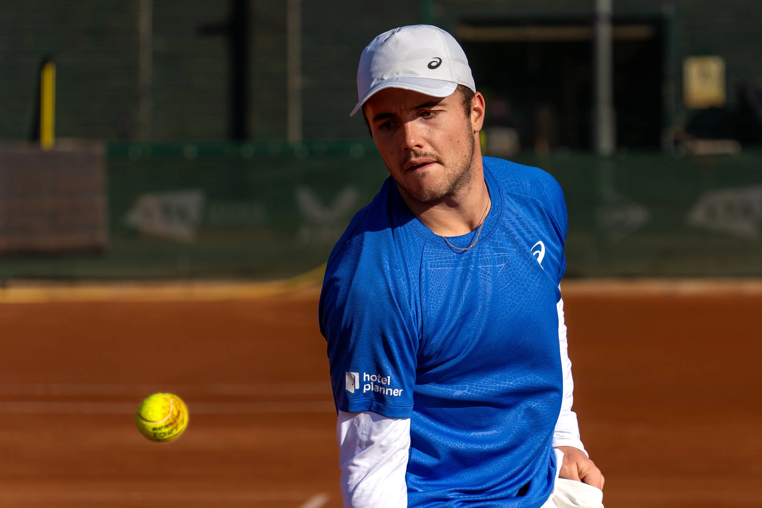 A male tennis player in a blue shirt and white hat preparing to hit a tennis ball on a clay court.