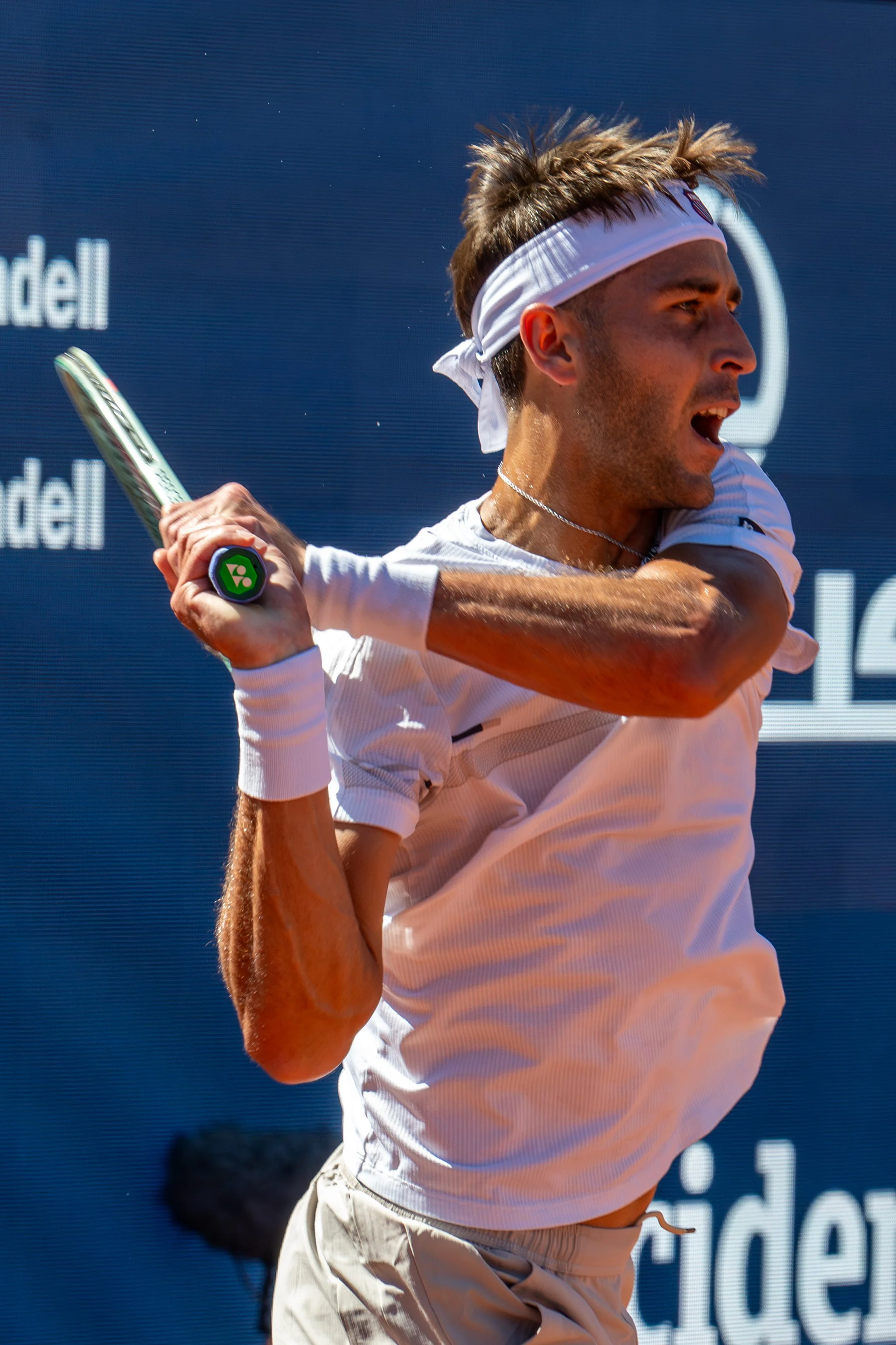 Tomas Martin Etcheverry in a white headband and white shirt prepares to hit a tennis ball with a racket, on an outdoor court.