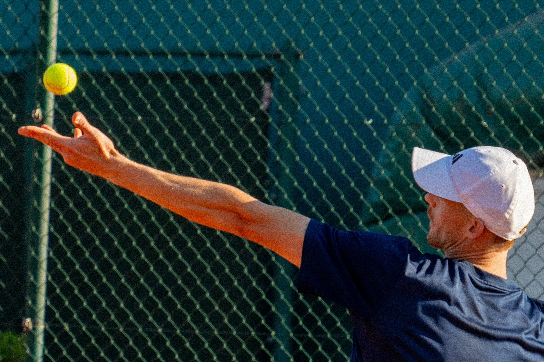 A man wearing a white baseball cap and navy shirt extending his arm to hit a bright yellow tennis ball on a tennis court with a green fence in the background.