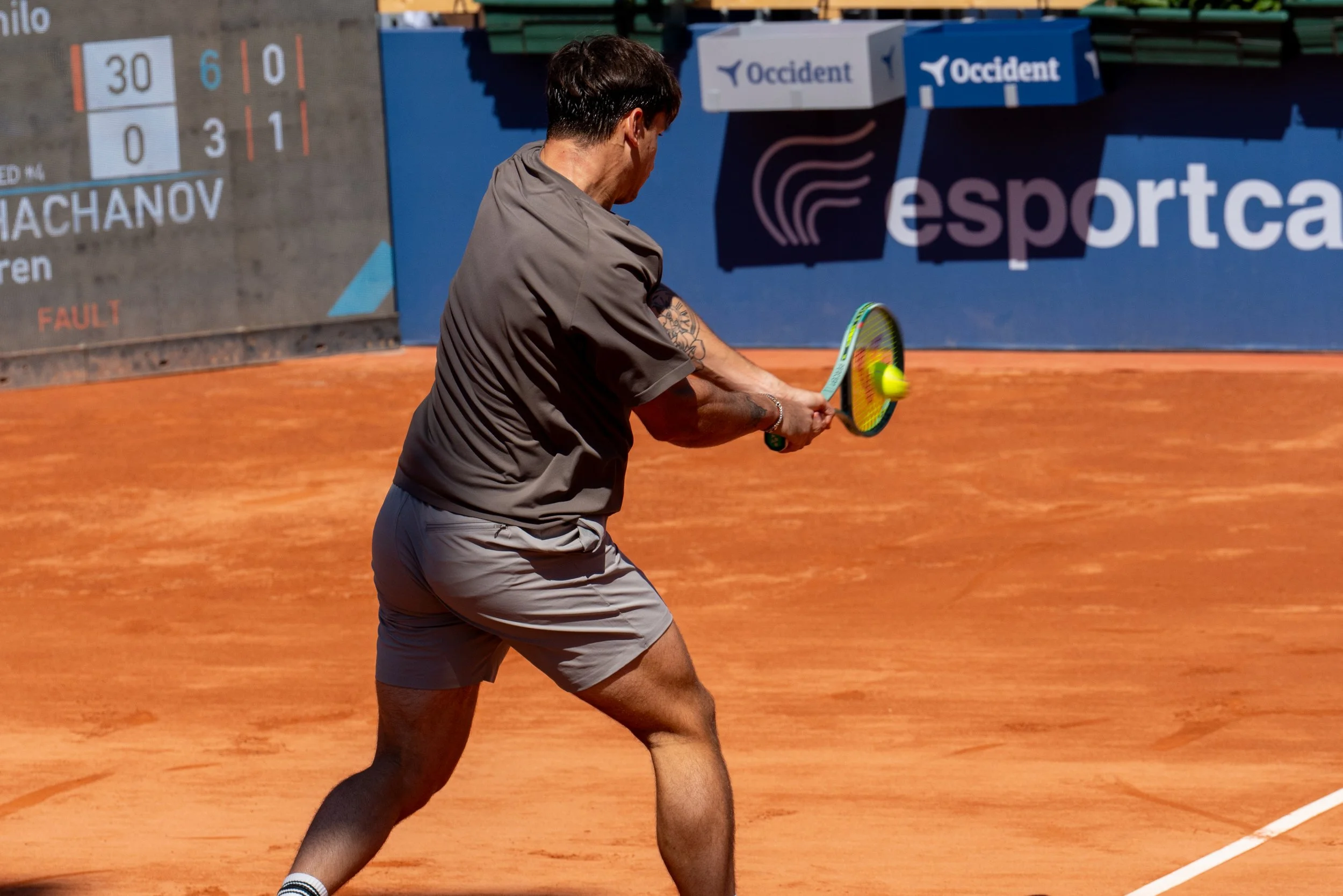 Camilo Ugo Carabelli on an orange clay court, wearing a gray shirt and shorts, hitting a tennis ball with a racket.