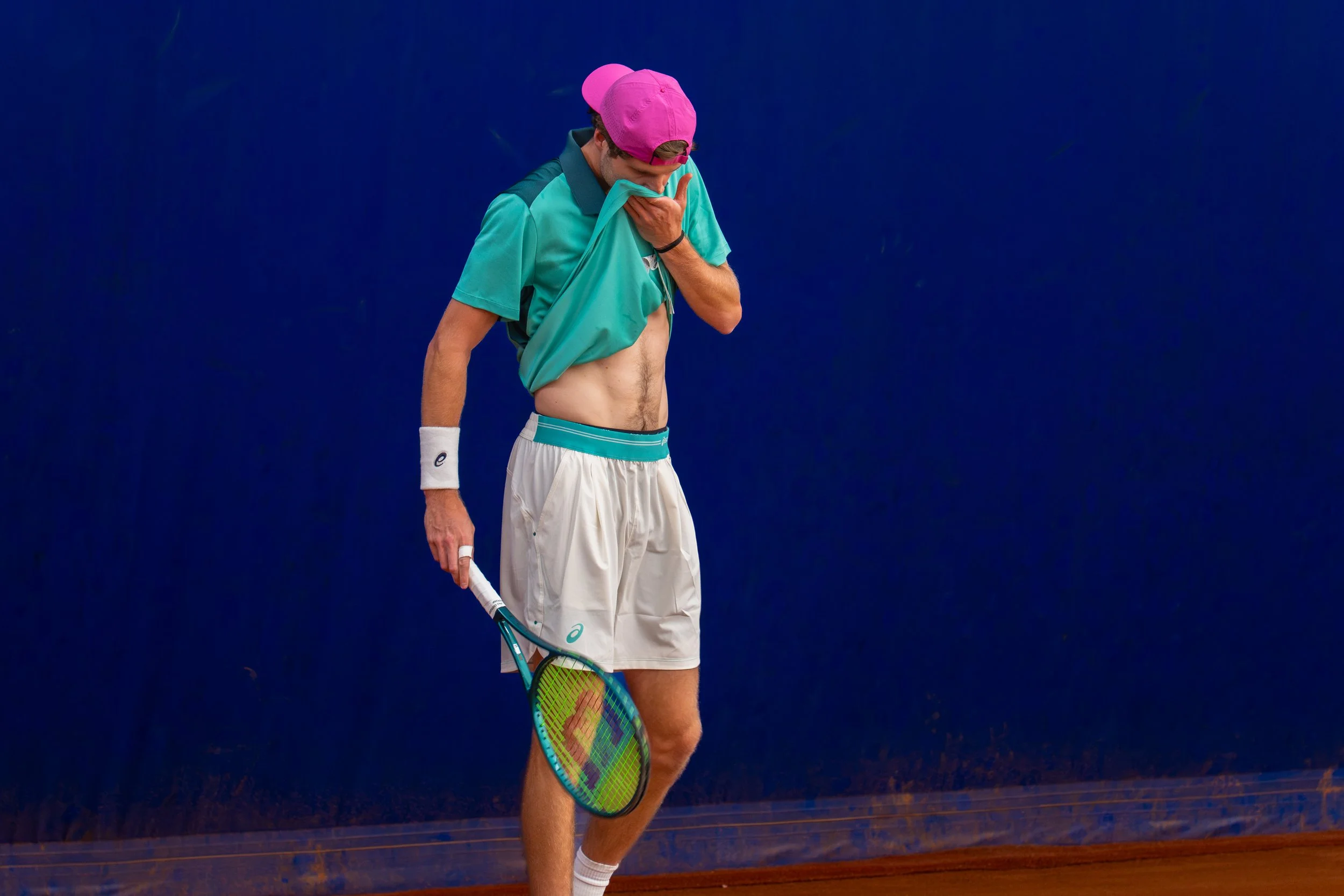 A male tennis player in a turquoise shirt, white shorts, and a pink cap stands on a tennis court, lifting his shirt to check his stomach while holding a tennis racket in his right hand.