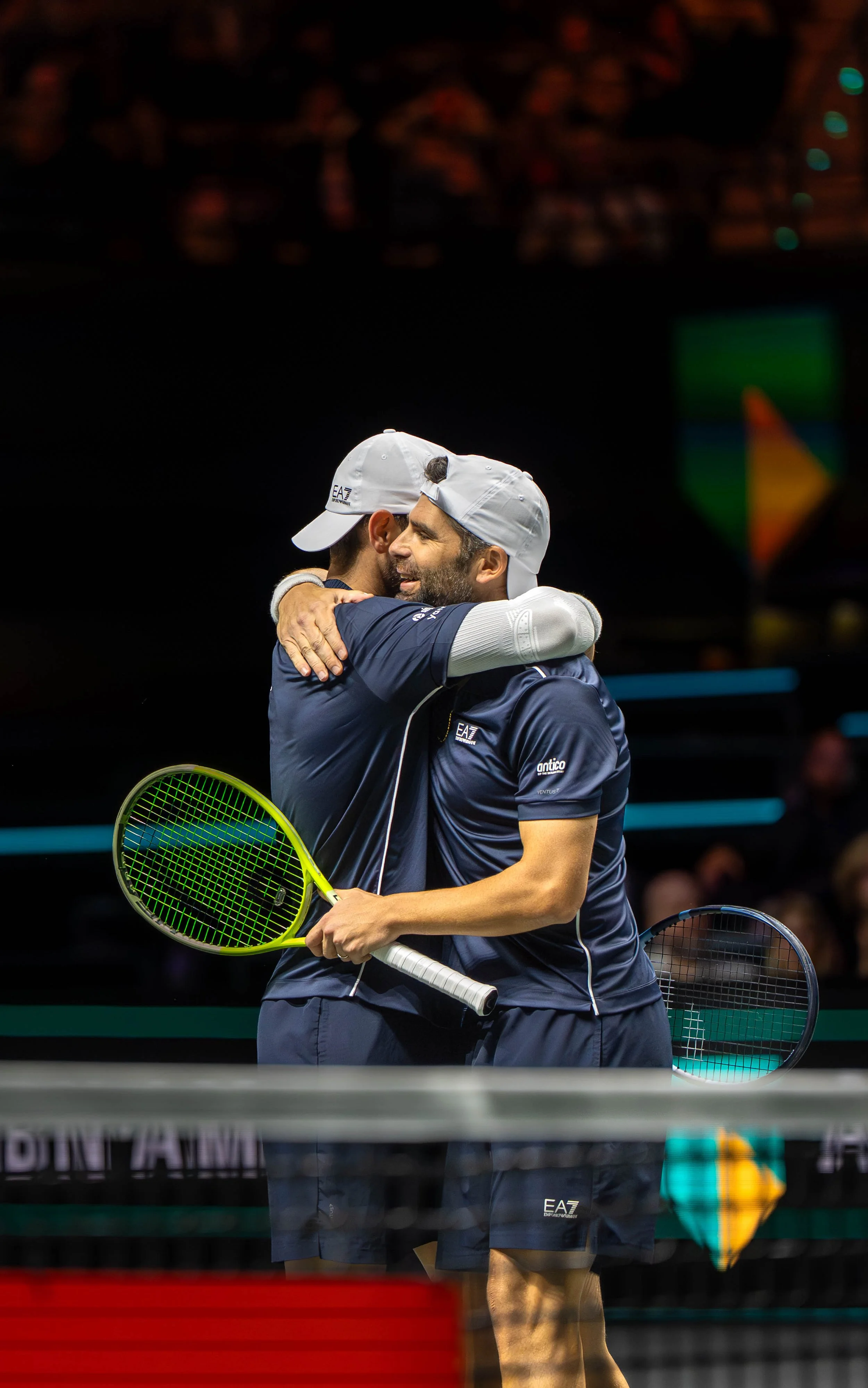 Two male tennis players wearing navy blue shirts and white caps hugging after a match on an indoor court.